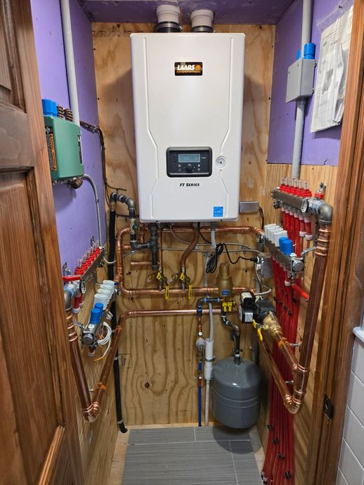 A Navien tankless water heater mounted on a plywood wall in a utility closet, flanked by red manifold plumbing systems.