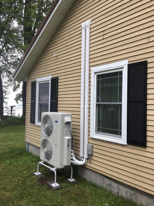 An outdoor HVAC heat pump unit mounted on a stand against a yellow-sided house with white vertical conduit pipe covers.