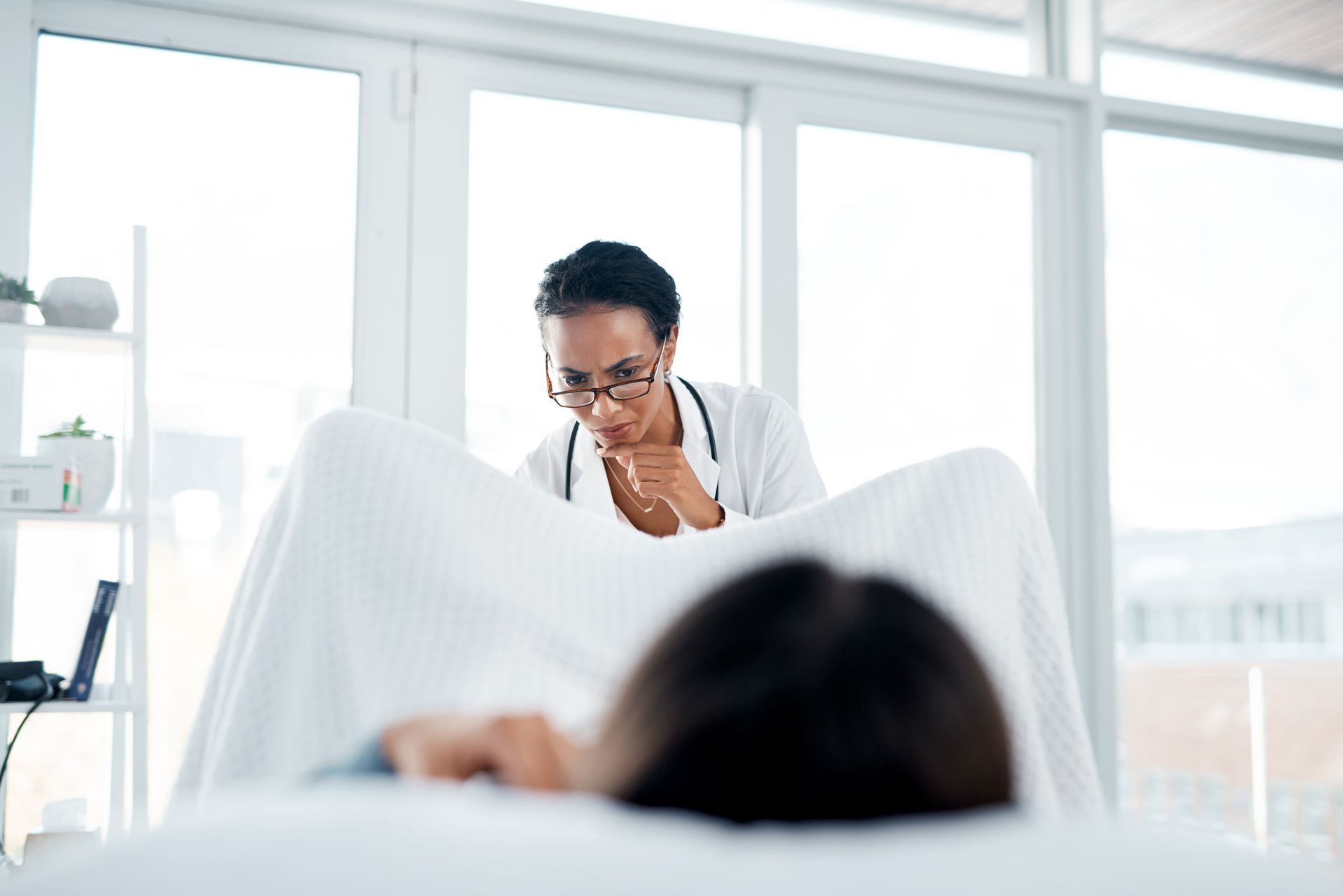 Female physician with glasses and stethoscope examining a patient lying on a clinic bed