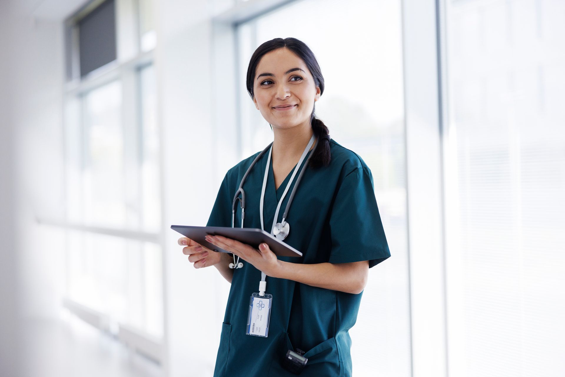 Healthcare professional in scrubs holding tablet in hospital corridor.