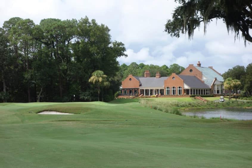 Golf course with a sand bunker and pond beside a large clubhouse under cloudy skies