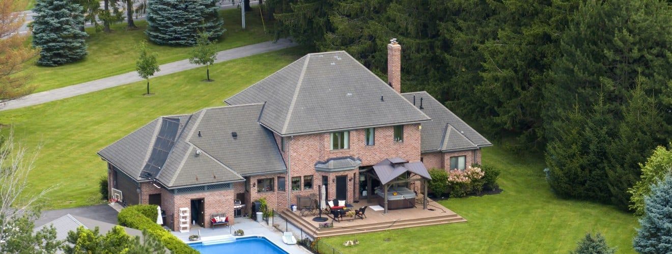 Aerial view of a large brick house with a gray roof surrounded by a green lawn and trees.