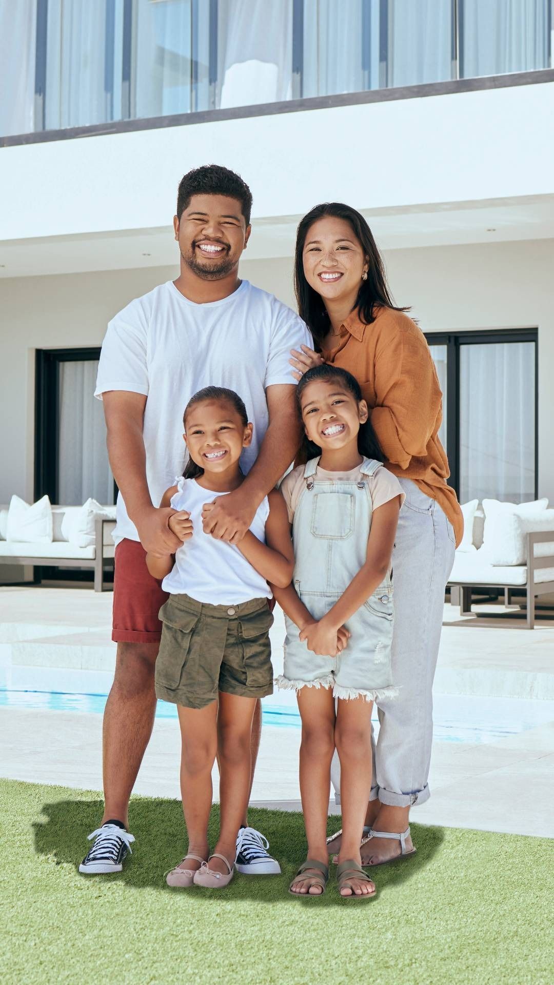 Family of four smiling on a patio outside a modern house, standing close together by a pool.