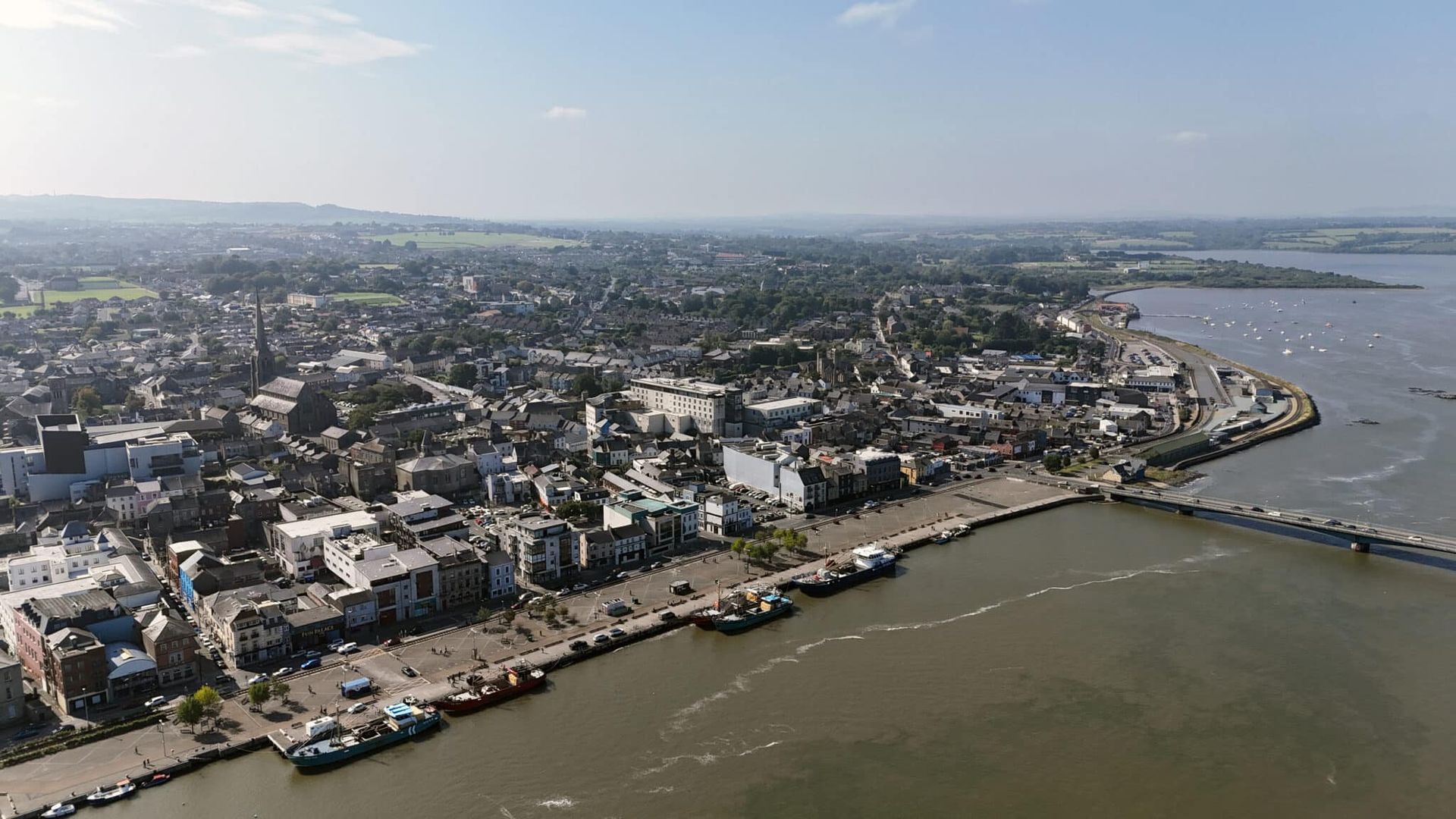 Aerial view of a coastal city with buildings, a waterfront road, and a broad bay under hazy skies