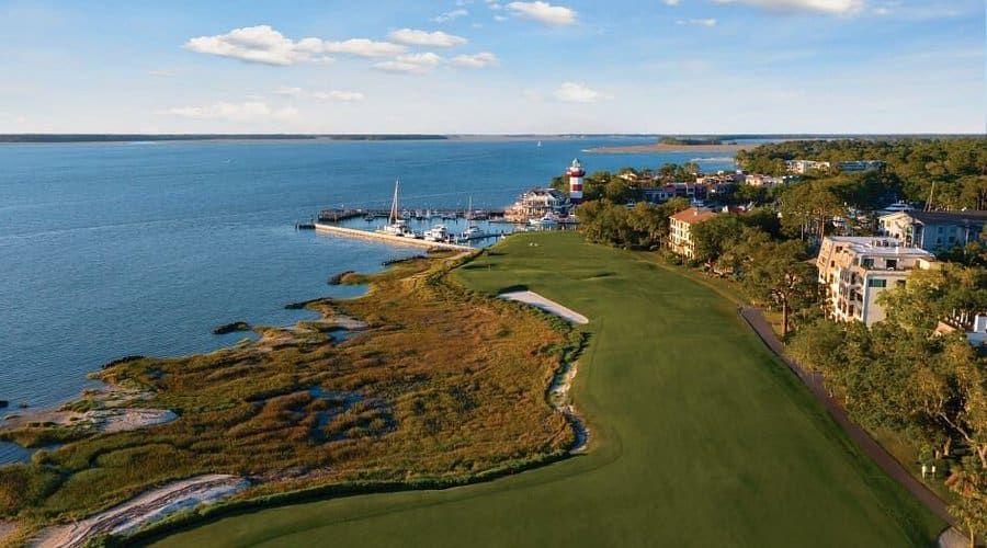 Coastal golf course beside calm blue water, with a marina and seaside homes under a partly cloudy sky