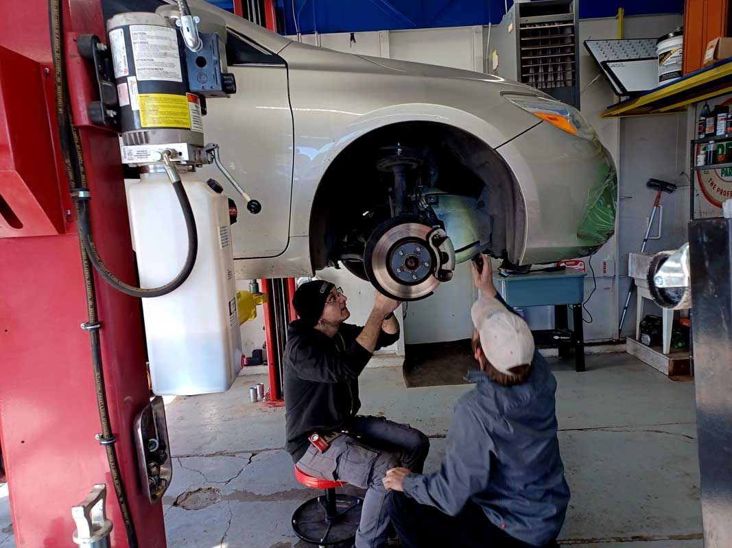 Two mechanics work under a silver car on a lift in an auto repair shop. | Milestone Auto Repair LLC