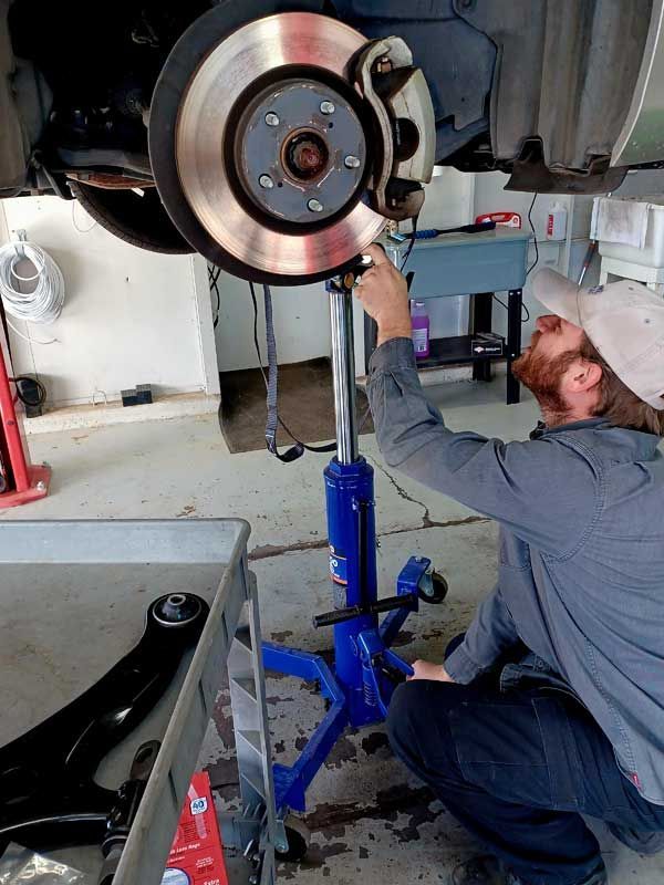 Mechanic inspecting a car’s brake disc while kneeling beside a raised vehicle in a garage | Milestone Auto Repair LLC