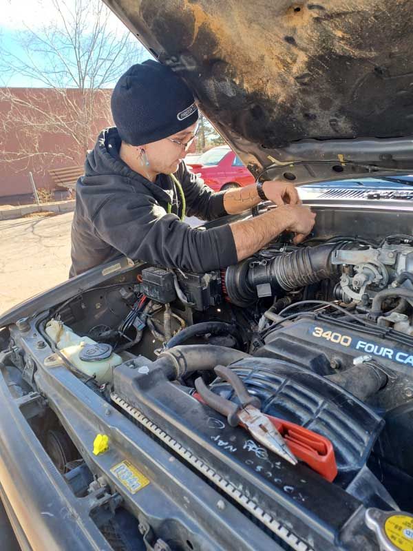 Mechanic working under a lifted car hood outdoors, repairing the engine bay with tools. | Milestone Auto Repair LLC