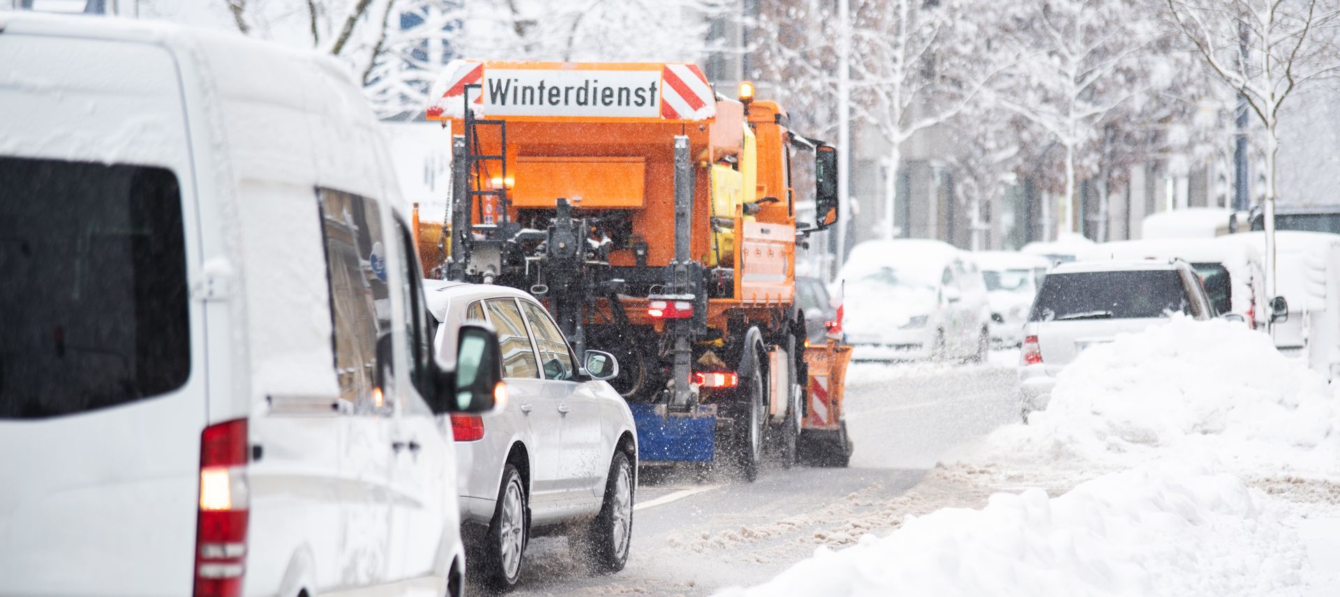 Ein Schneepflug fährt neben einem weißen Lieferwagen eine schneebedeckte Straße entlang.