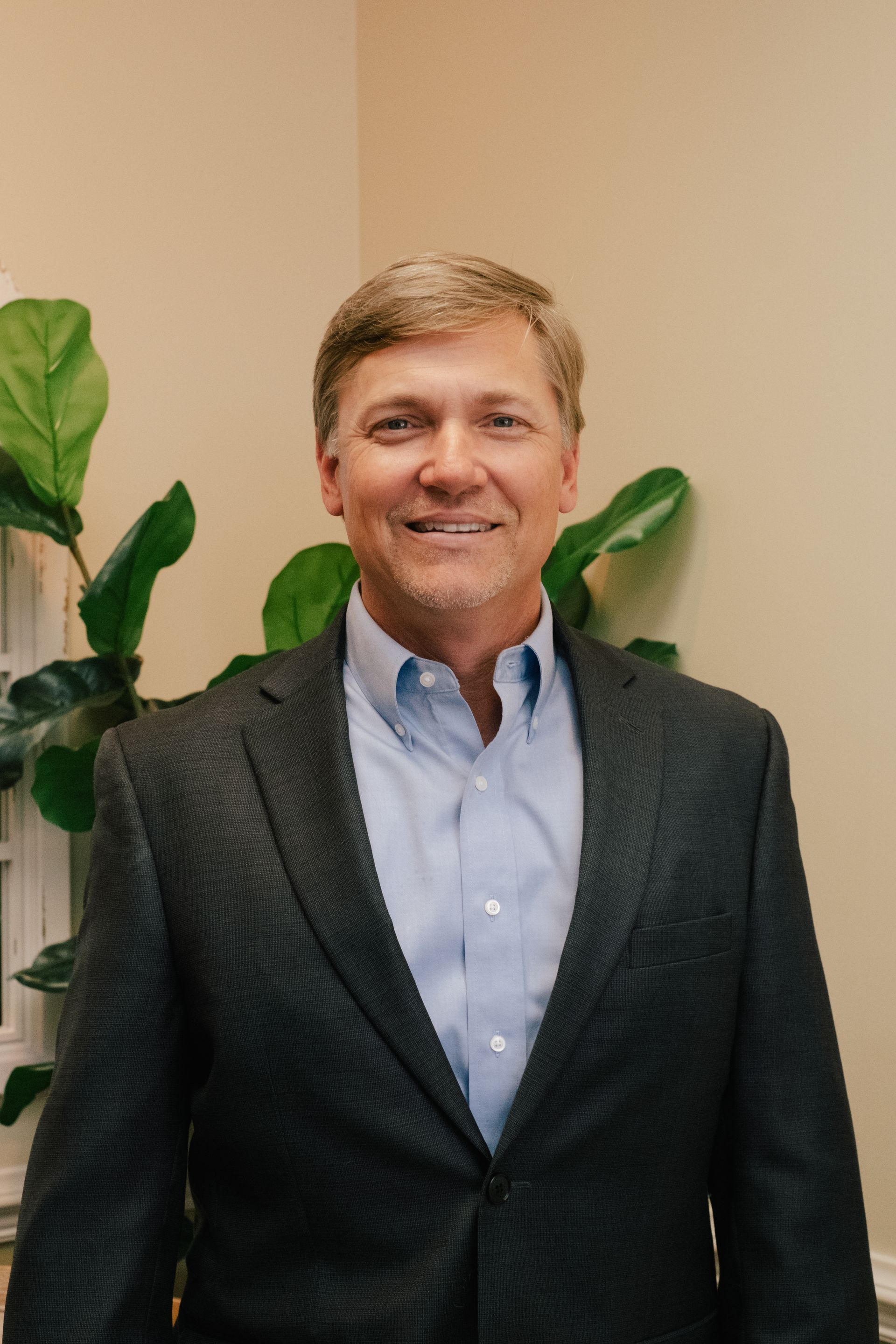 A man in a suit and blue shirt is standing in front of a plant.