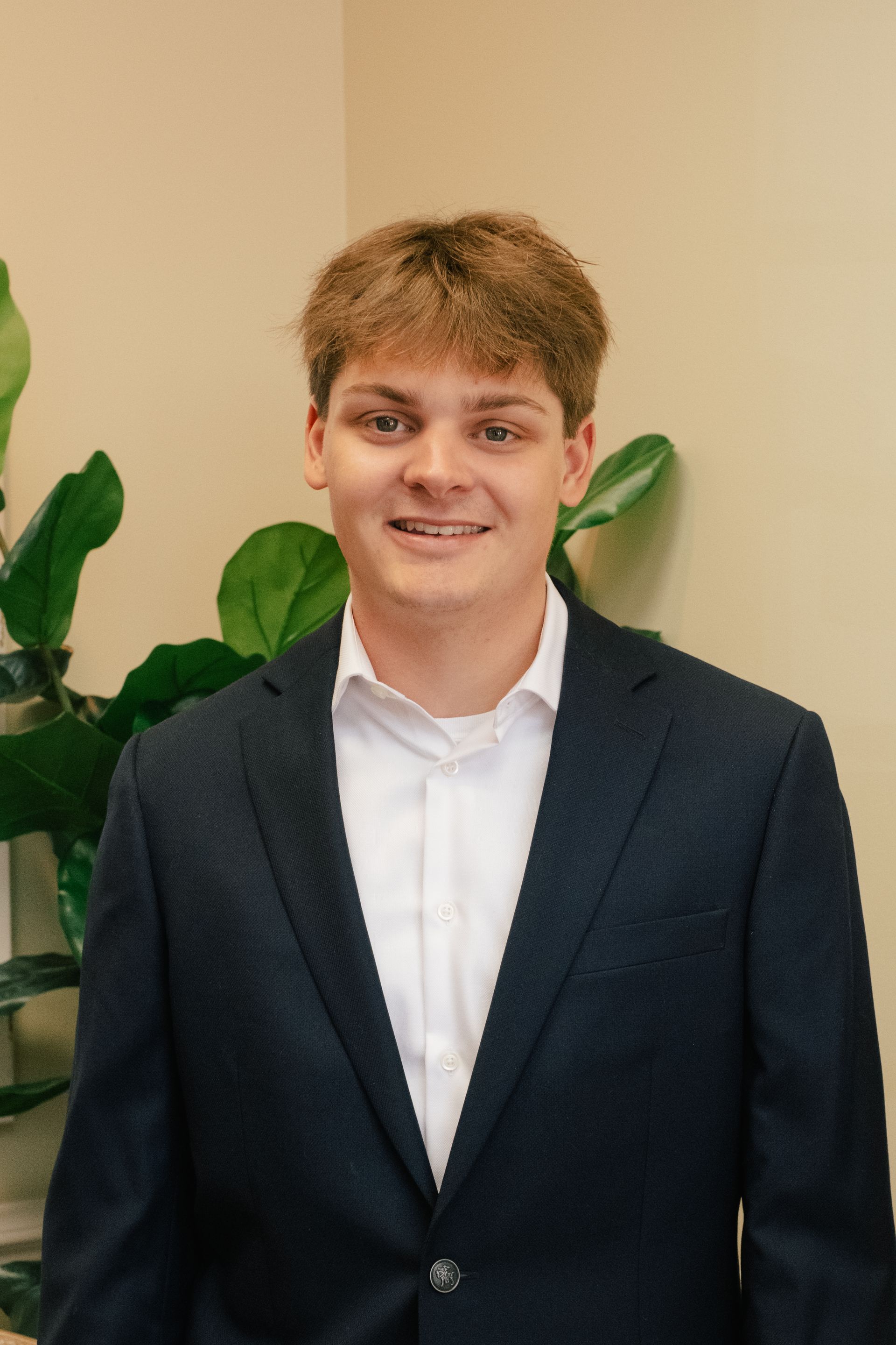 A young man in a suit and white shirt is standing in front of a plant.