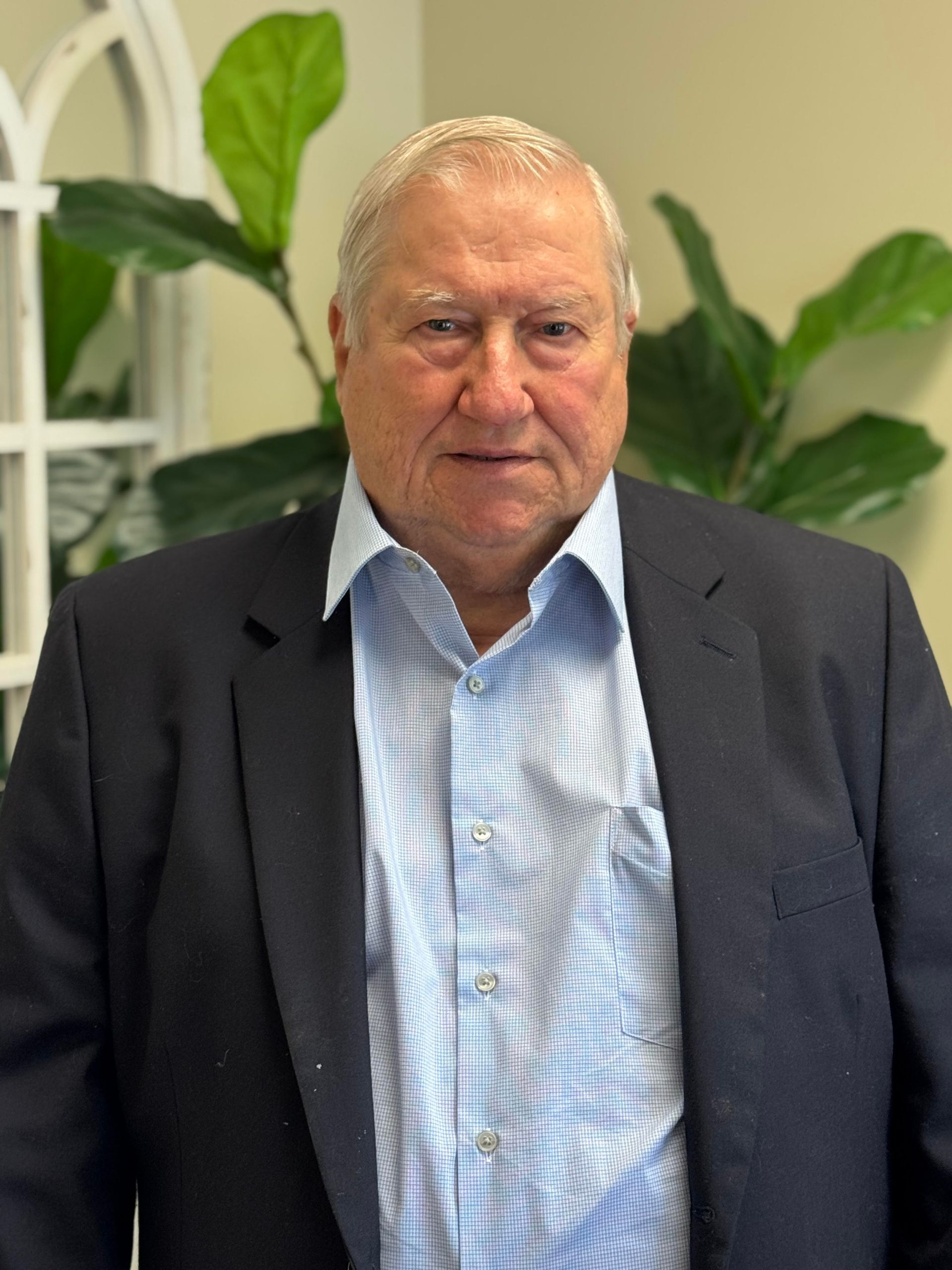 A man in a suit and blue shirt is standing in front of a plant.