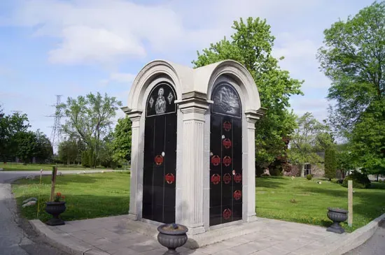 Small ornate mausoleum with black doors and red wreaths in a grassy cemetery