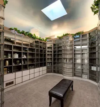 Curved room lined with floor-to-ceiling shelves and a black bench under a skylight, with plants along the top.