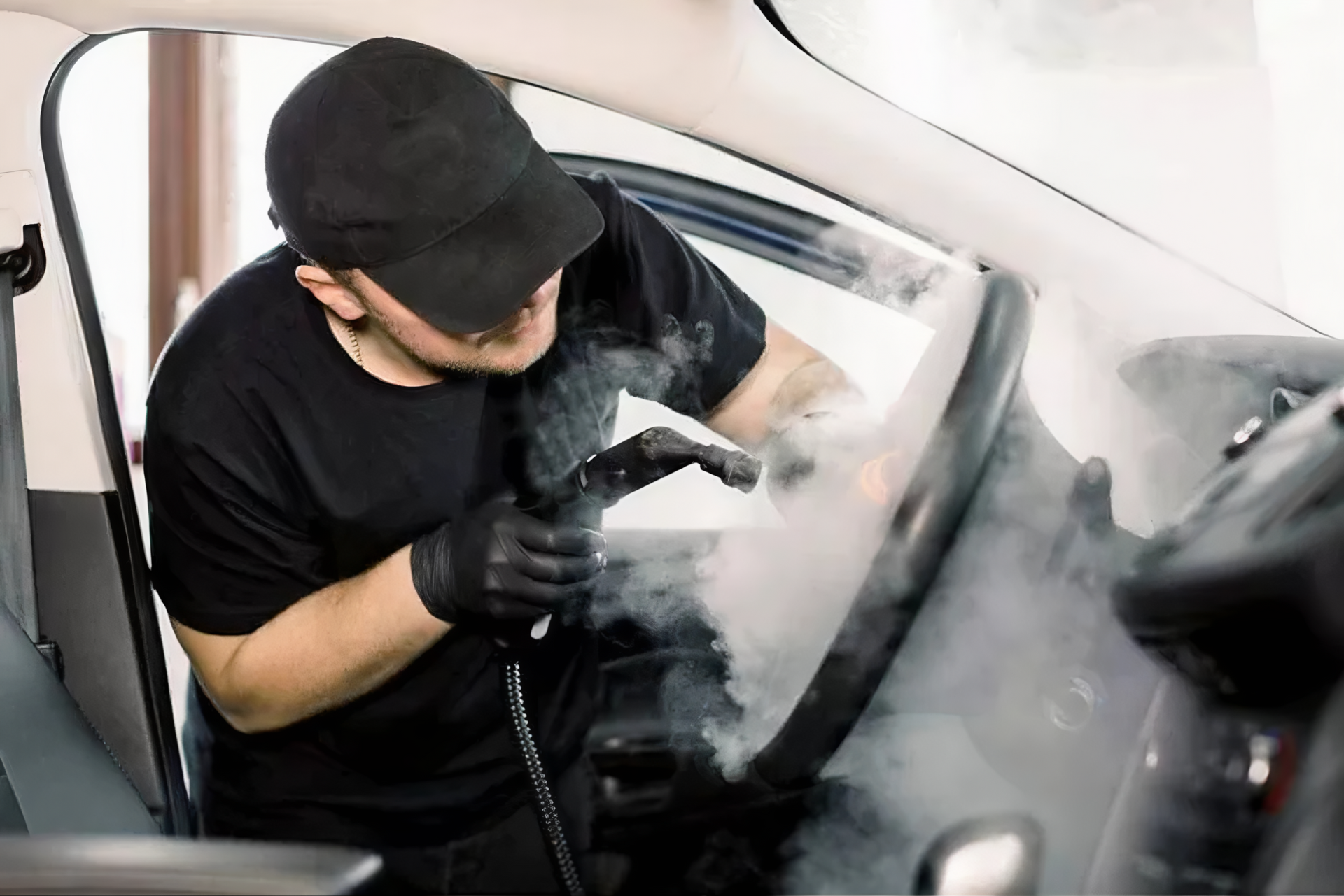 Person in black cleaning a car interior with a steam cleaner, visible steam.