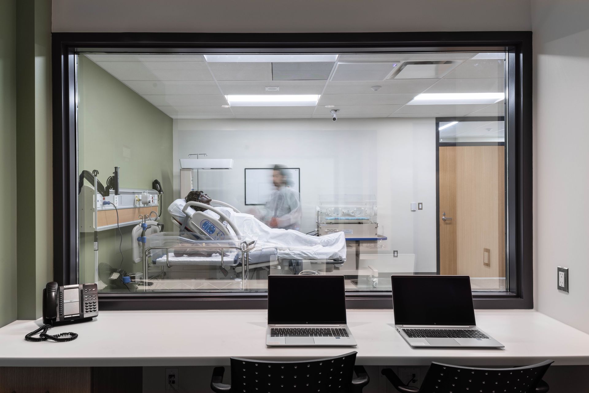 A medical training room with a patient in bed, observed by others, viewed through a large window from a workstation with laptops.