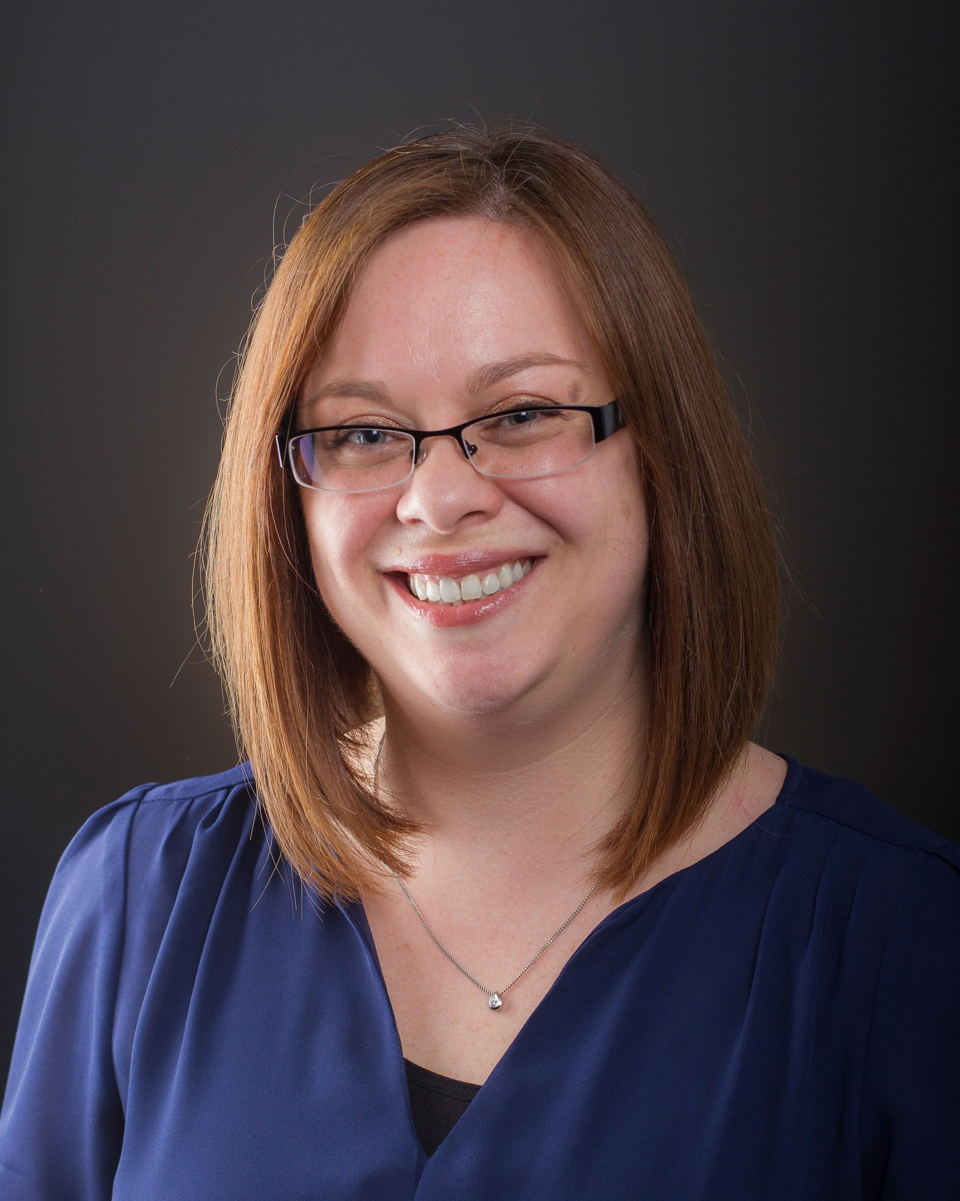 Woman with glasses smiling, wearing a blue blouse, against a dark gray background.