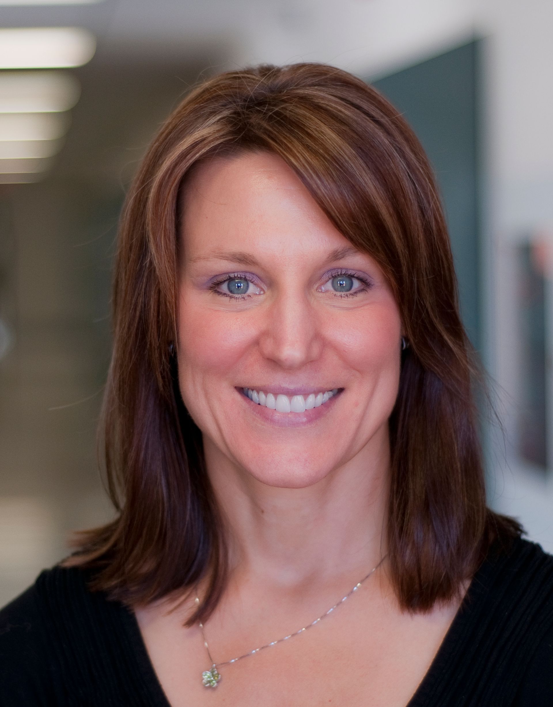 Woman with brown hair smiles at the camera, wearing a black shirt and necklace.