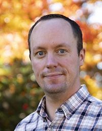 Man in plaid shirt smiles in front of fall foliage.