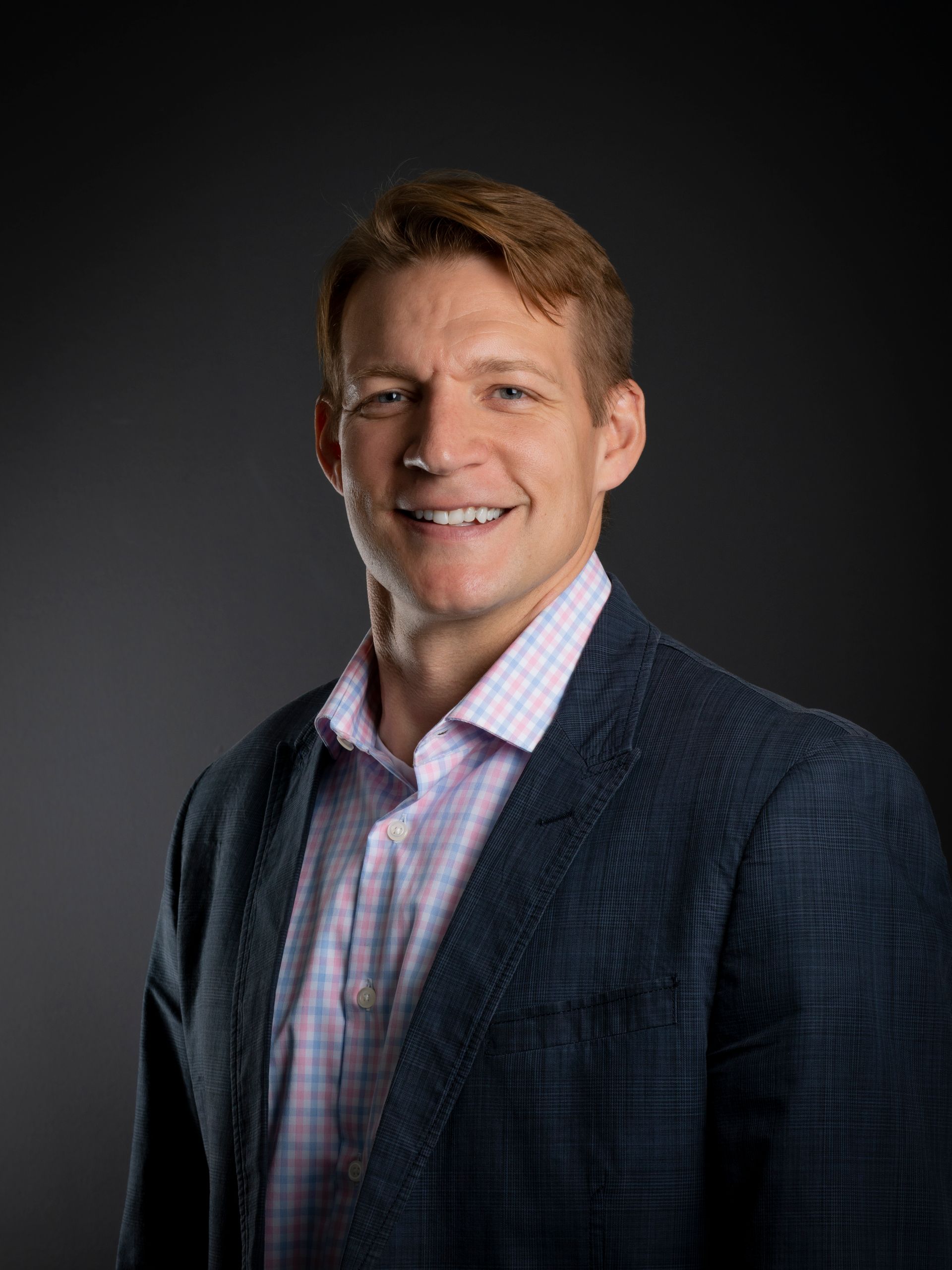 Man in blazer smiling, indoors against a gray background.