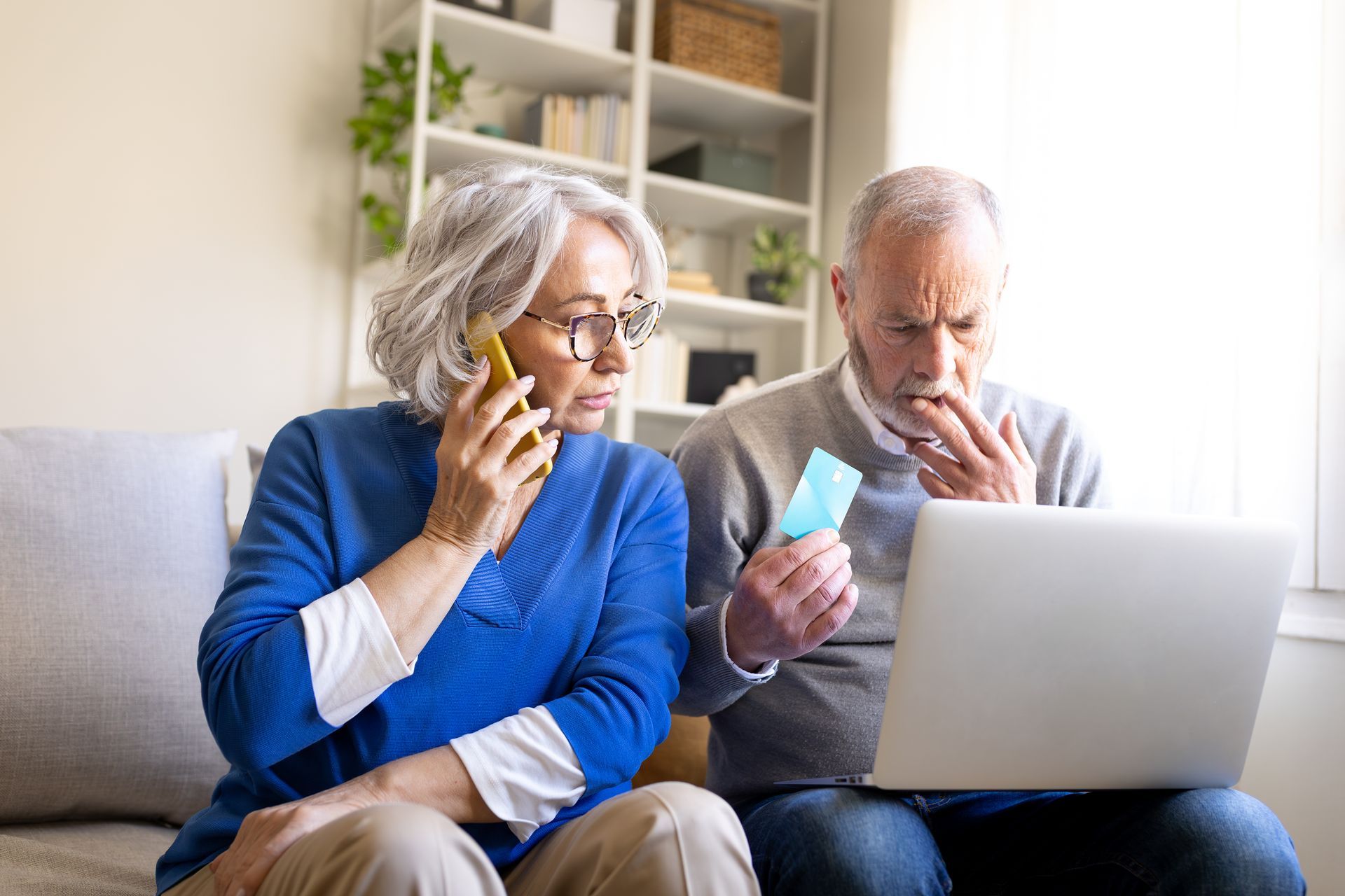Two older adults on a couch reviewing a laptop and a credit card in a bright living room