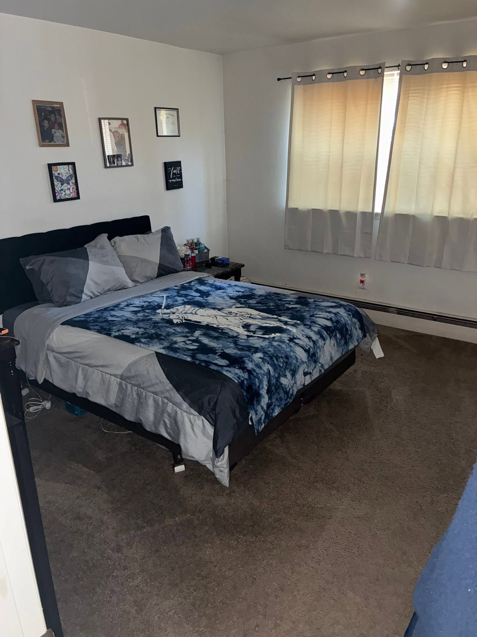 Bedroom with a bed, blue-gray bedding, wall photos, and a window with beige curtains.