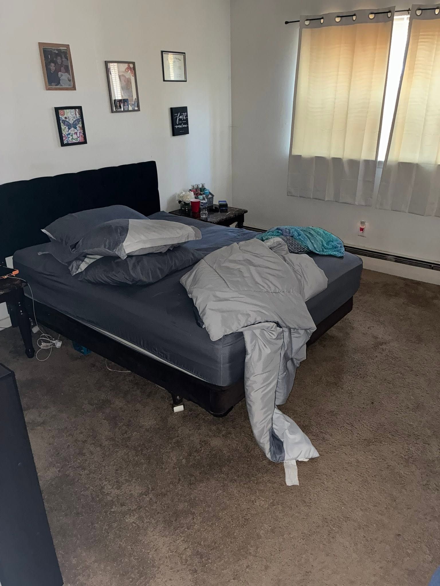 Unmade bed in a carpeted bedroom with a window, wall photos, and clothes draped over the bed.