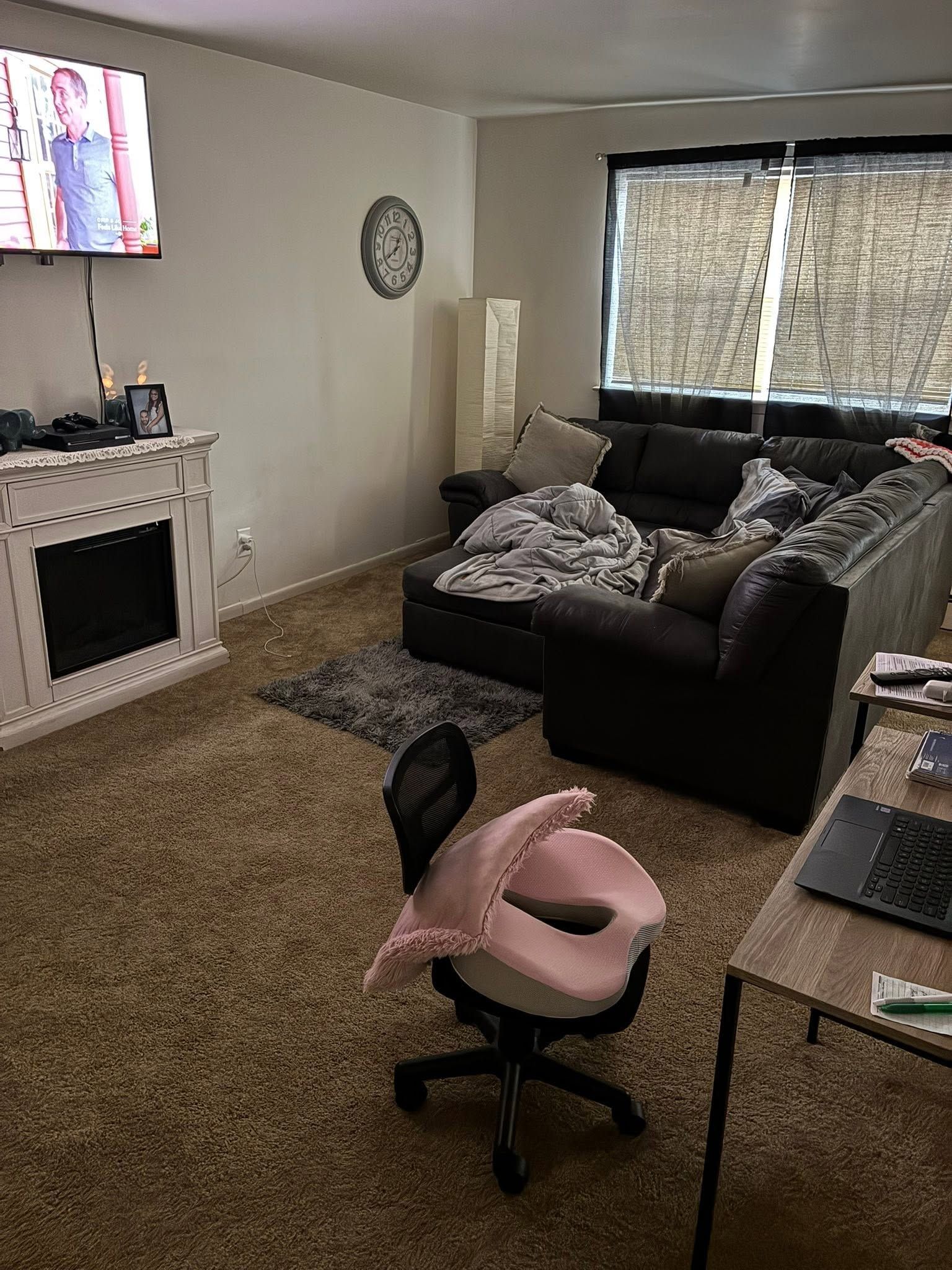 Living room with black sofa, pink office chair, TV above fireplace, and patterned curtains on a window