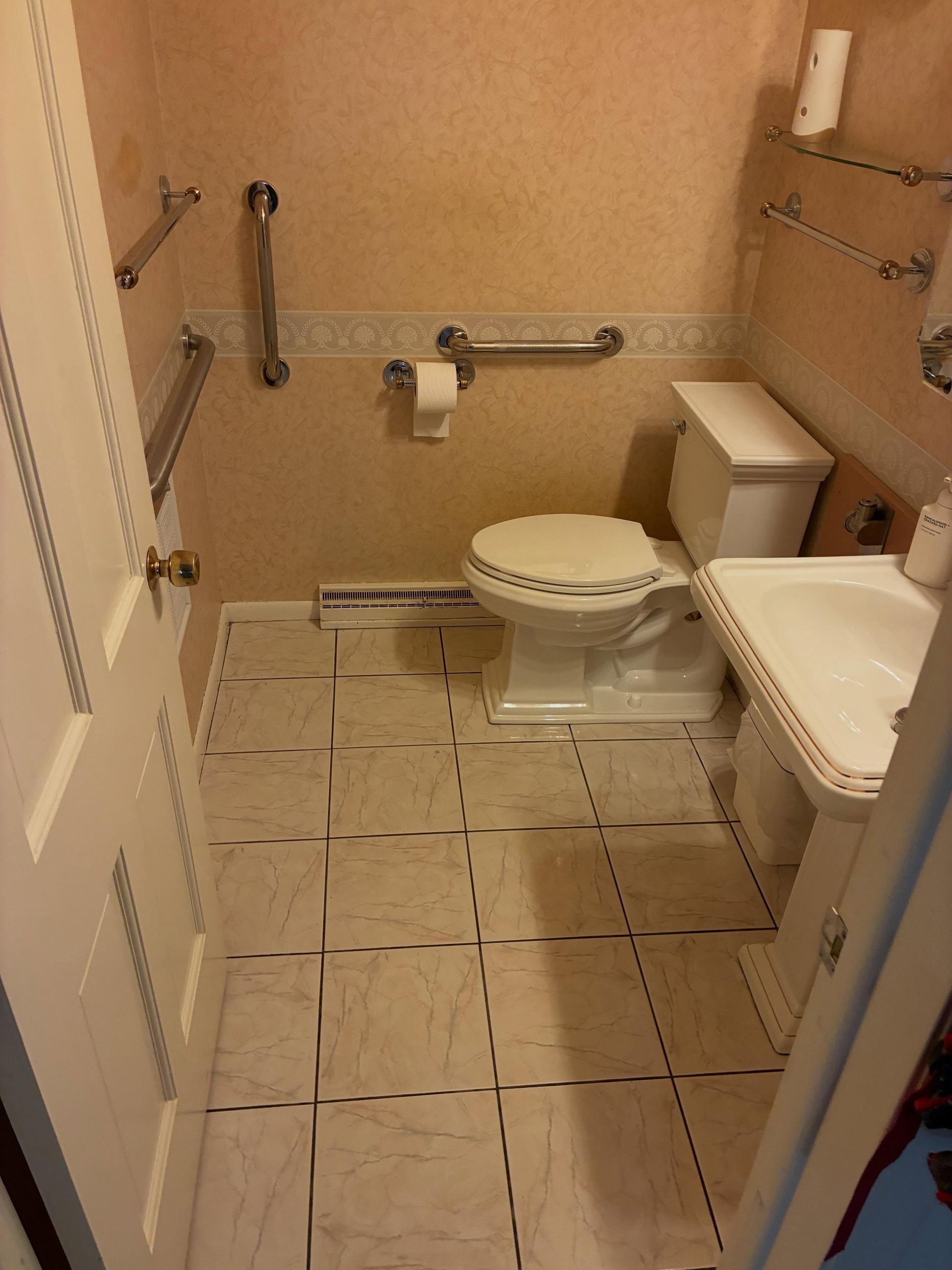 Small accessible bathroom with raised toilet, grab bars, wall sink, and tiled floor.
