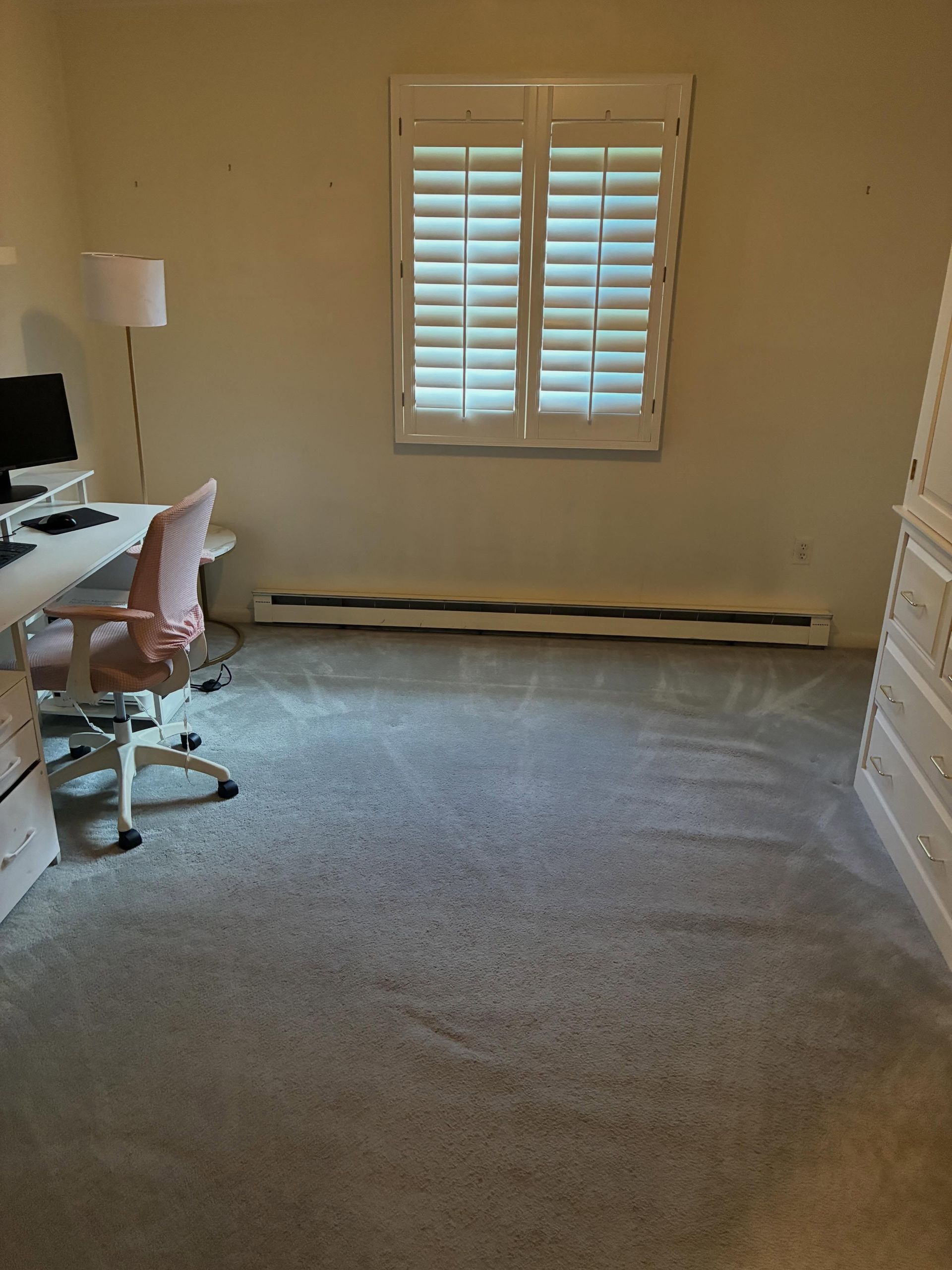 Small home office with desk, pink chair, window shutters, and carpeted floor.