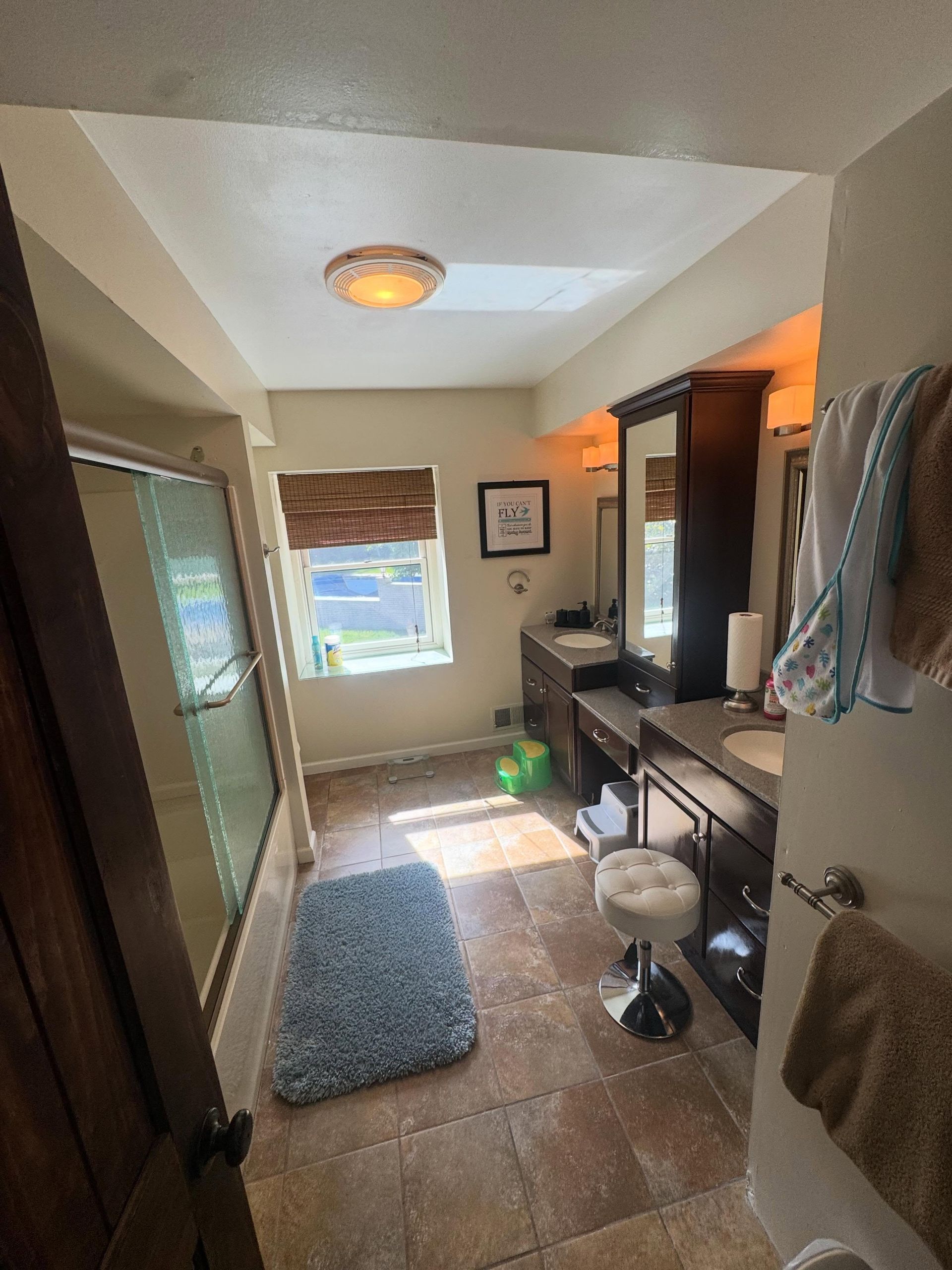 Narrow bathroom with beige tile, blue rug, vanity sink, mirror, window, and towels hanging on the wall.