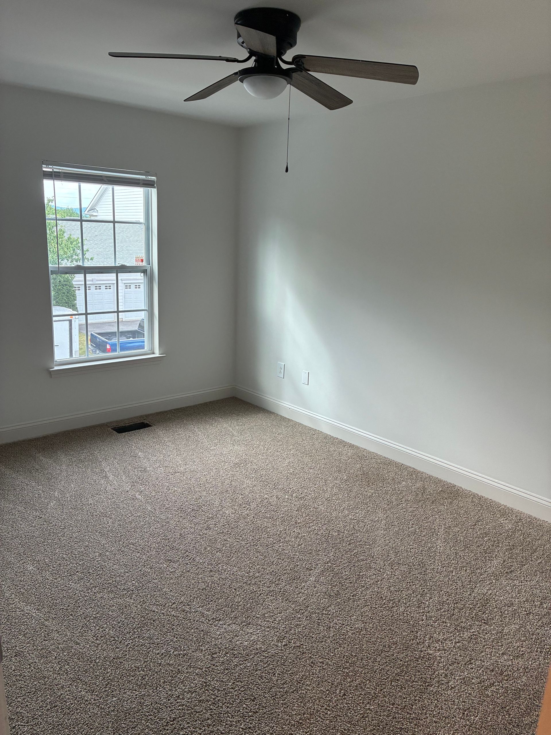 Empty carpeted bedroom with white walls, a ceiling fan, and a window letting in daylight.