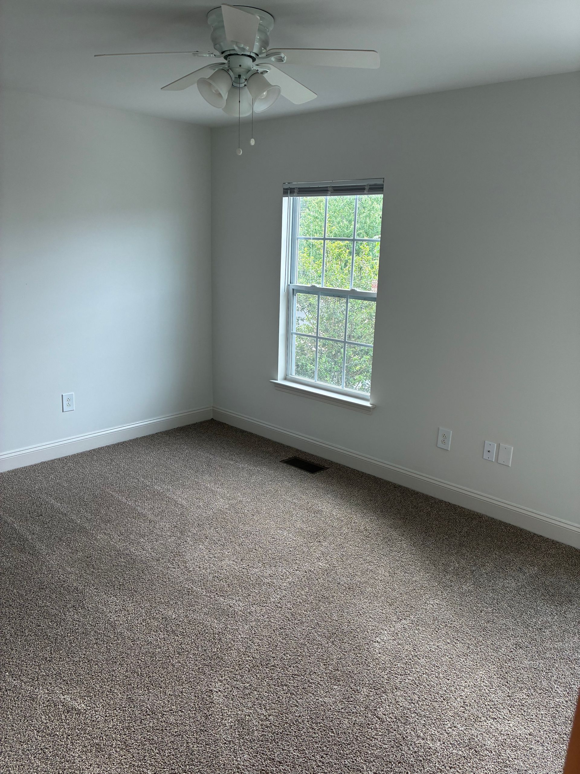 Empty carpeted bedroom with white walls, a ceiling fan, and a window letting in daylight.