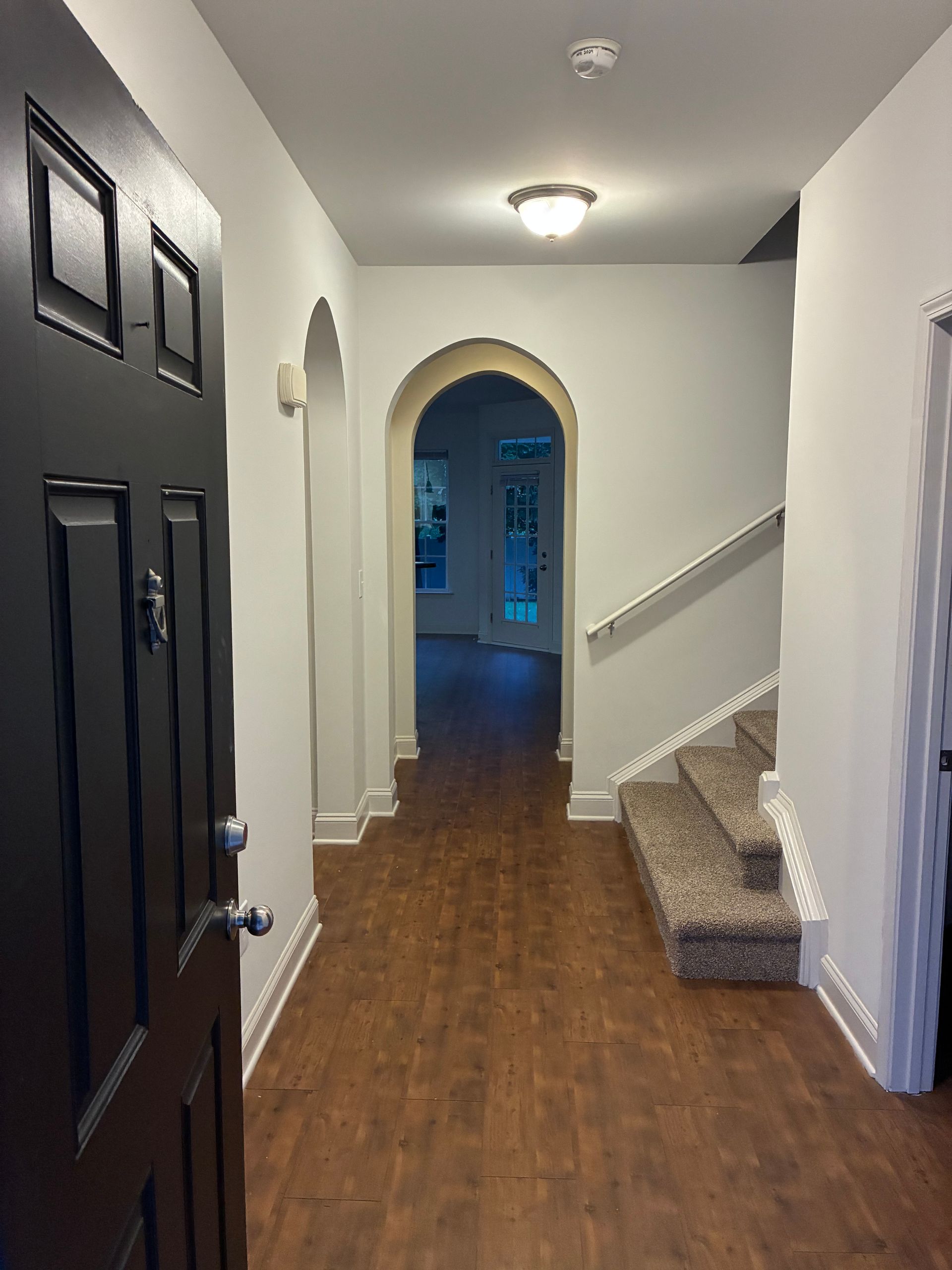 Narrow hallway with dark front door, arched doorway, stairs, and wood floor in a home
