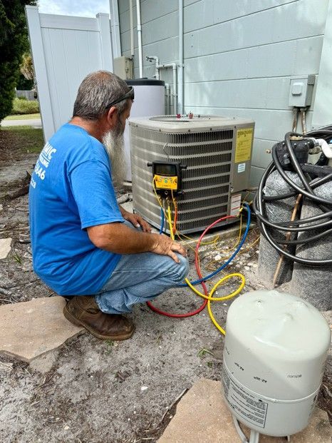 HVAC technician servicing an air conditioner outdoors, using gauges. Yellow, blue, and red hoses connected.