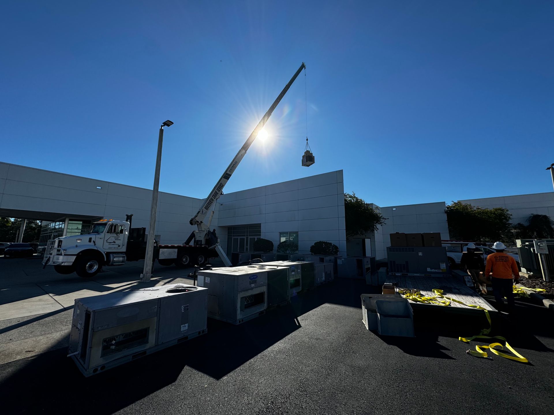 A crane lifts an object above a building on a sunny day. Workers and equipment are on site.