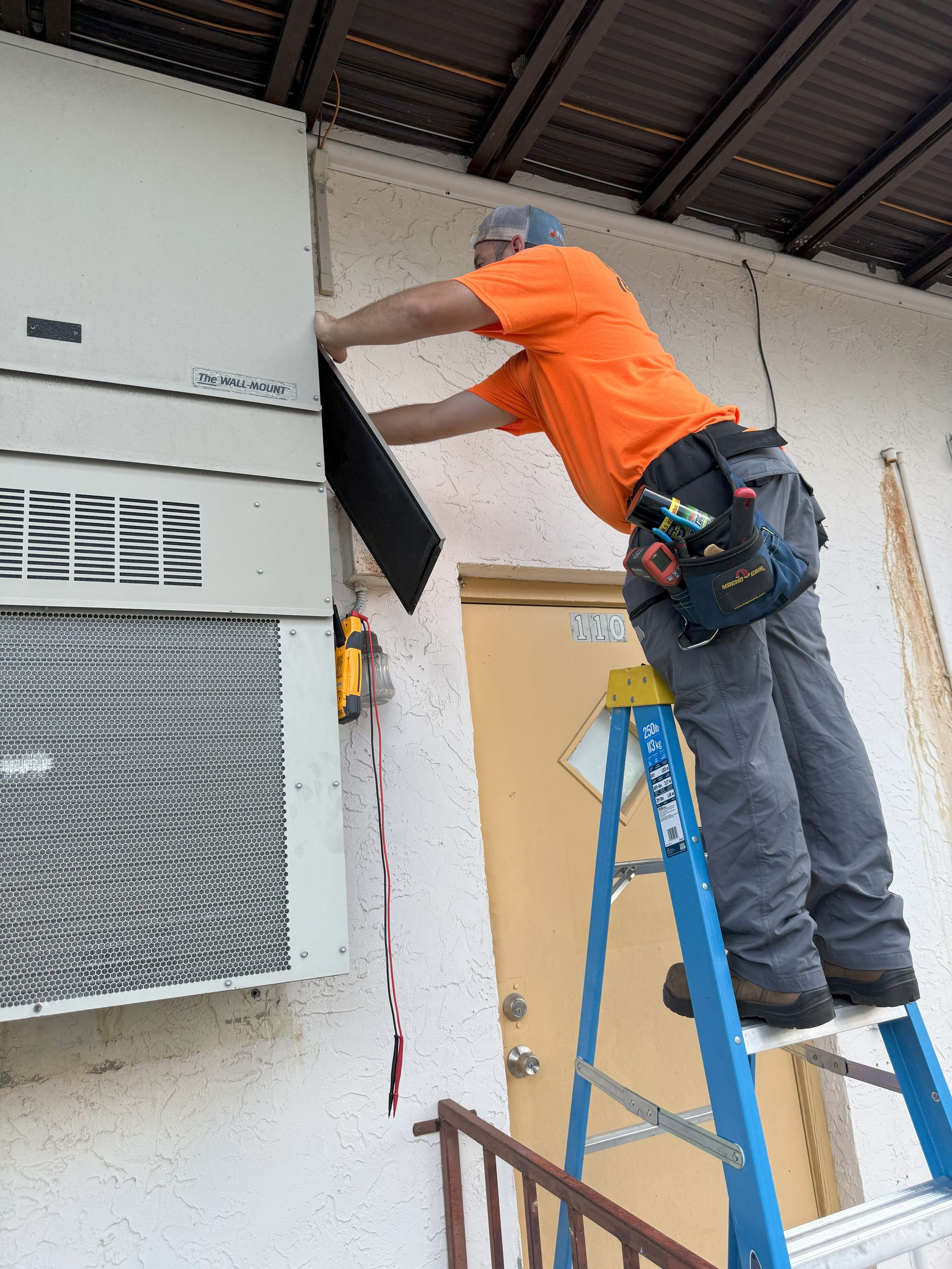 Man in orange shirt on ladder installing panel on building's exterior, near air conditioner.