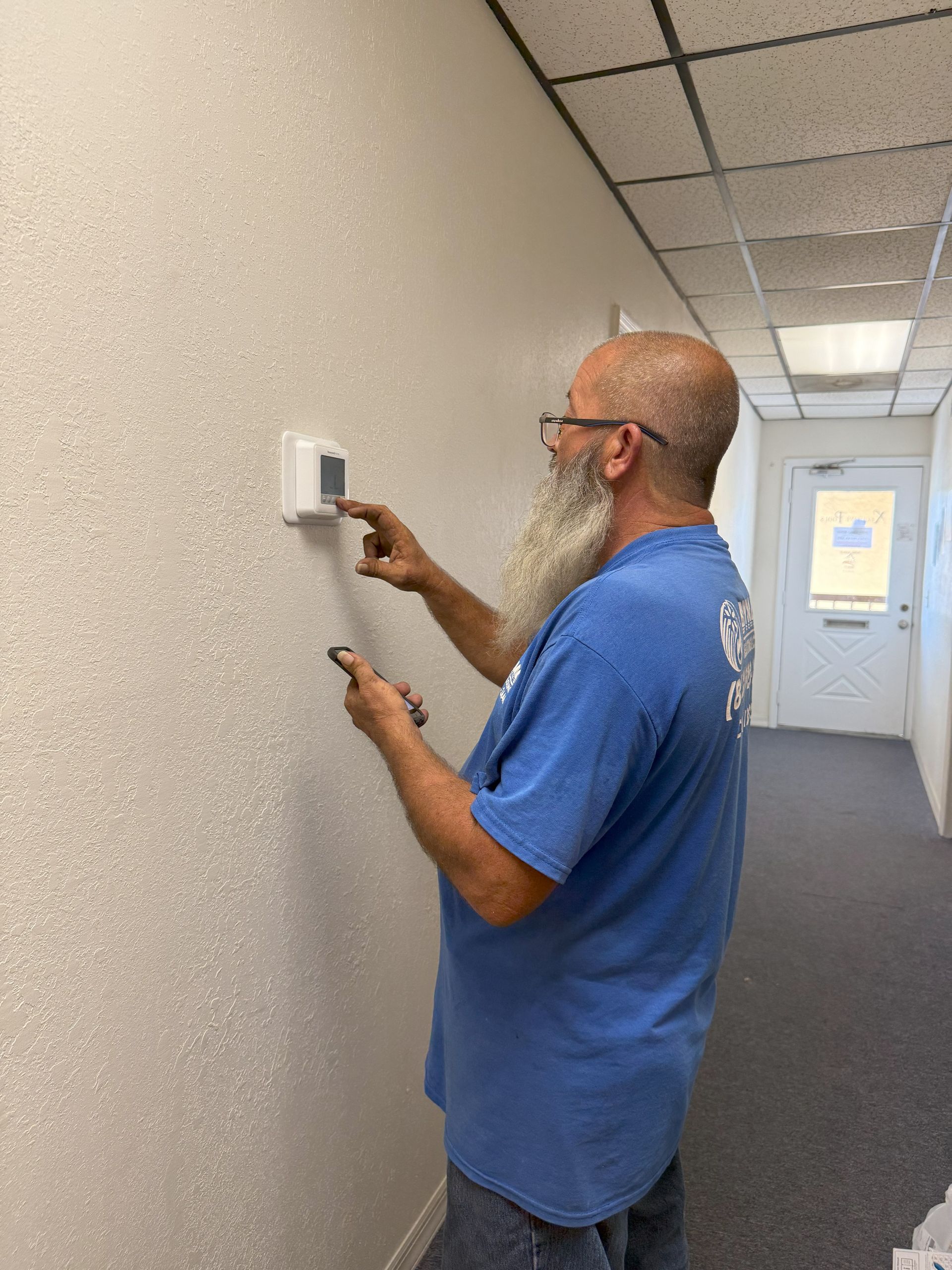Man adjusting thermostat on textured wall in office hallway. He has a beard and glasses, wearing blue shirt.