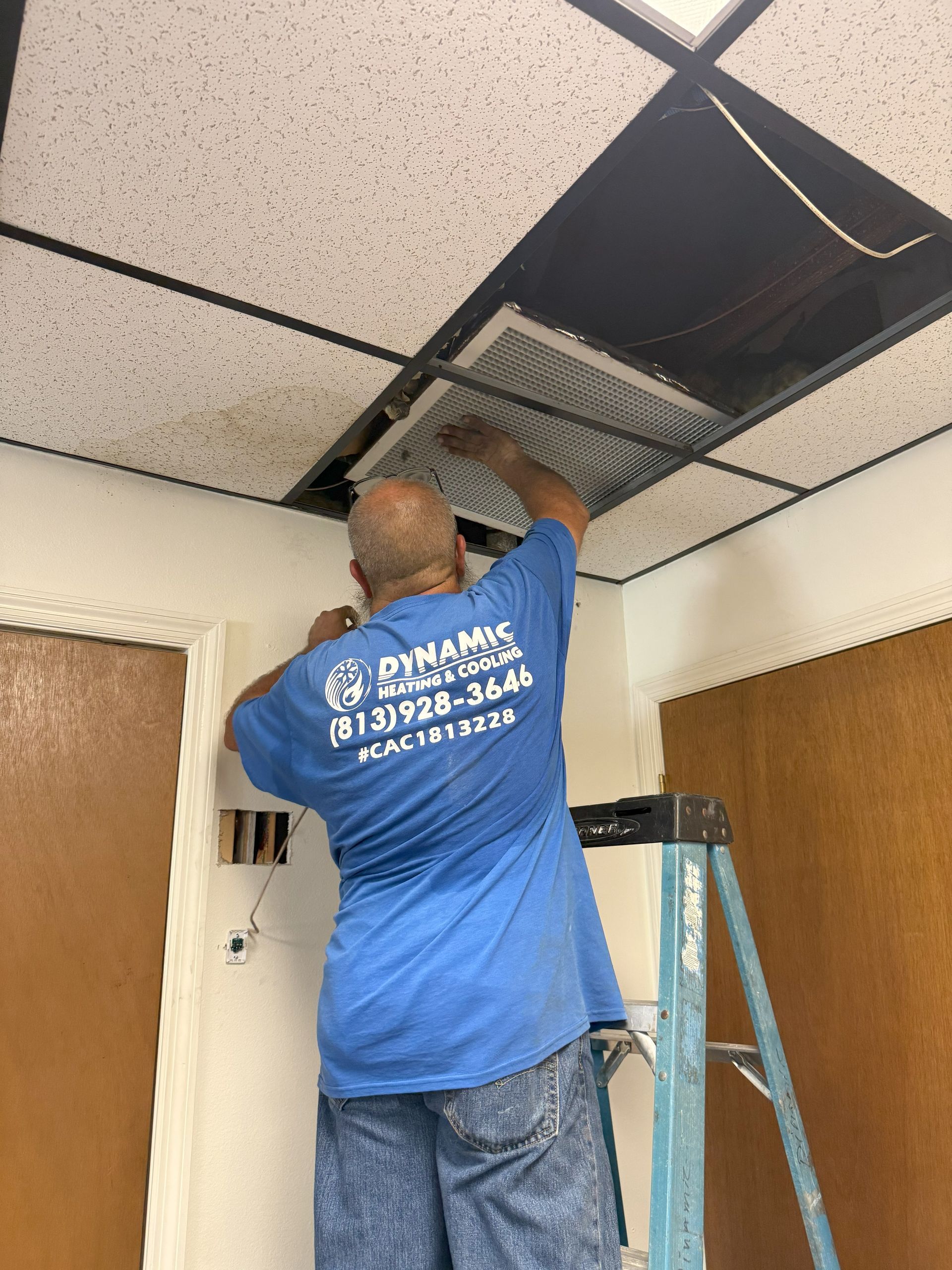 Man in blue shirt on a ladder, replacing a ceiling air vent in a room with brown doors and walls.
