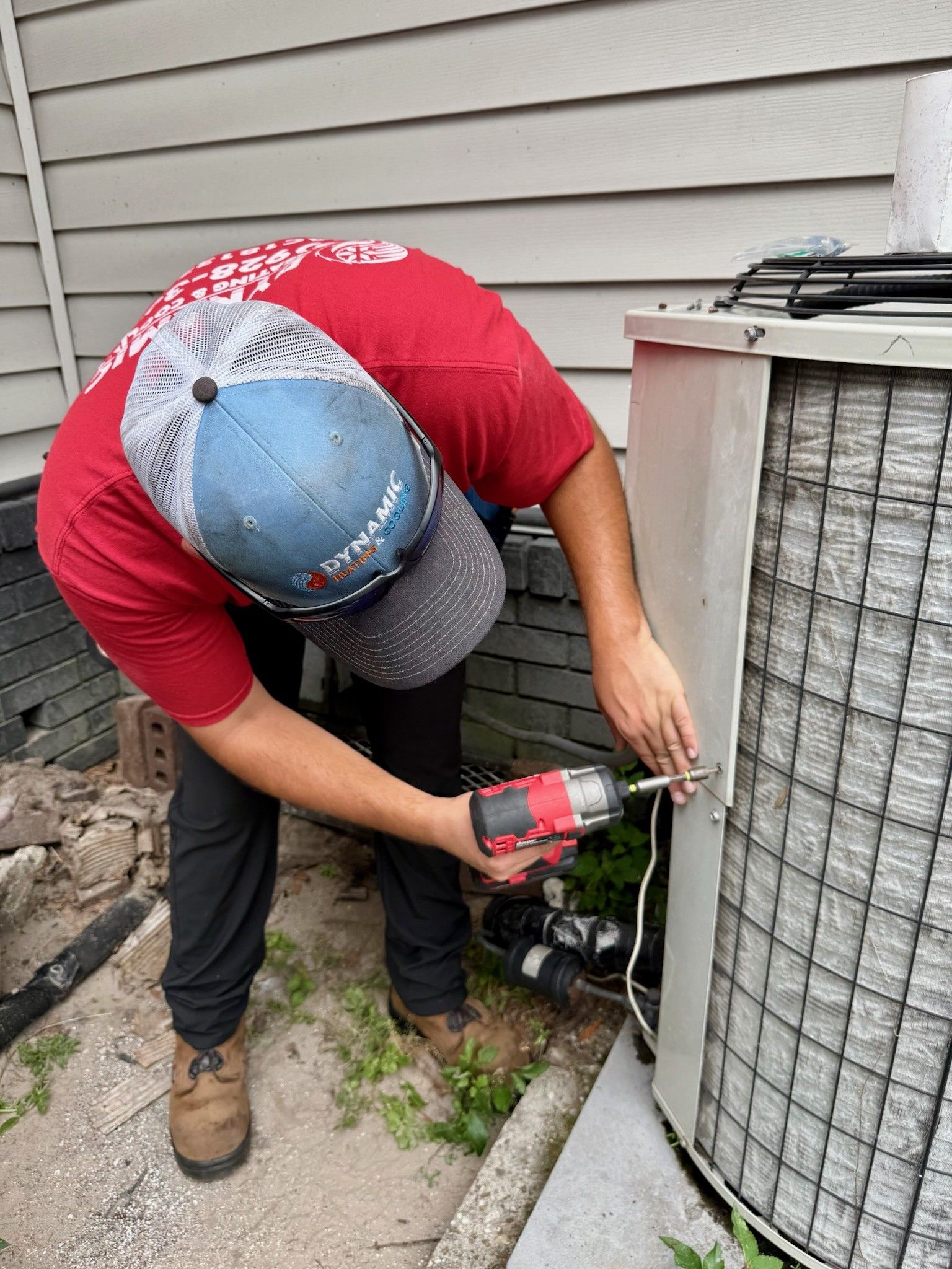 Man in red shirt fixing an air conditioning unit with a drill outdoors.