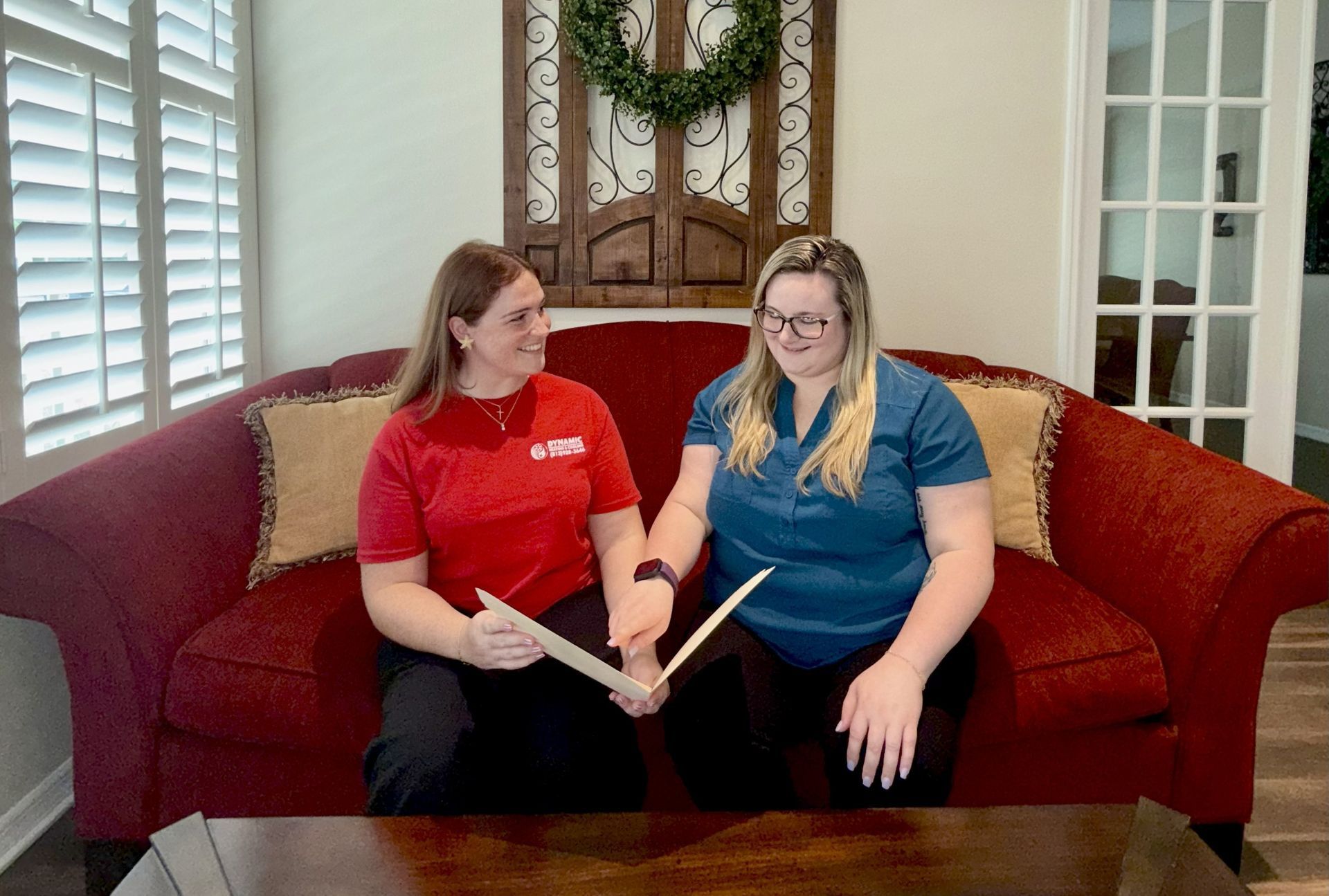 Two women, one in red and one in blue, seated on a red couch, holding and examining bamboo sticks.  They are smiling indoors with a wreath in the background.