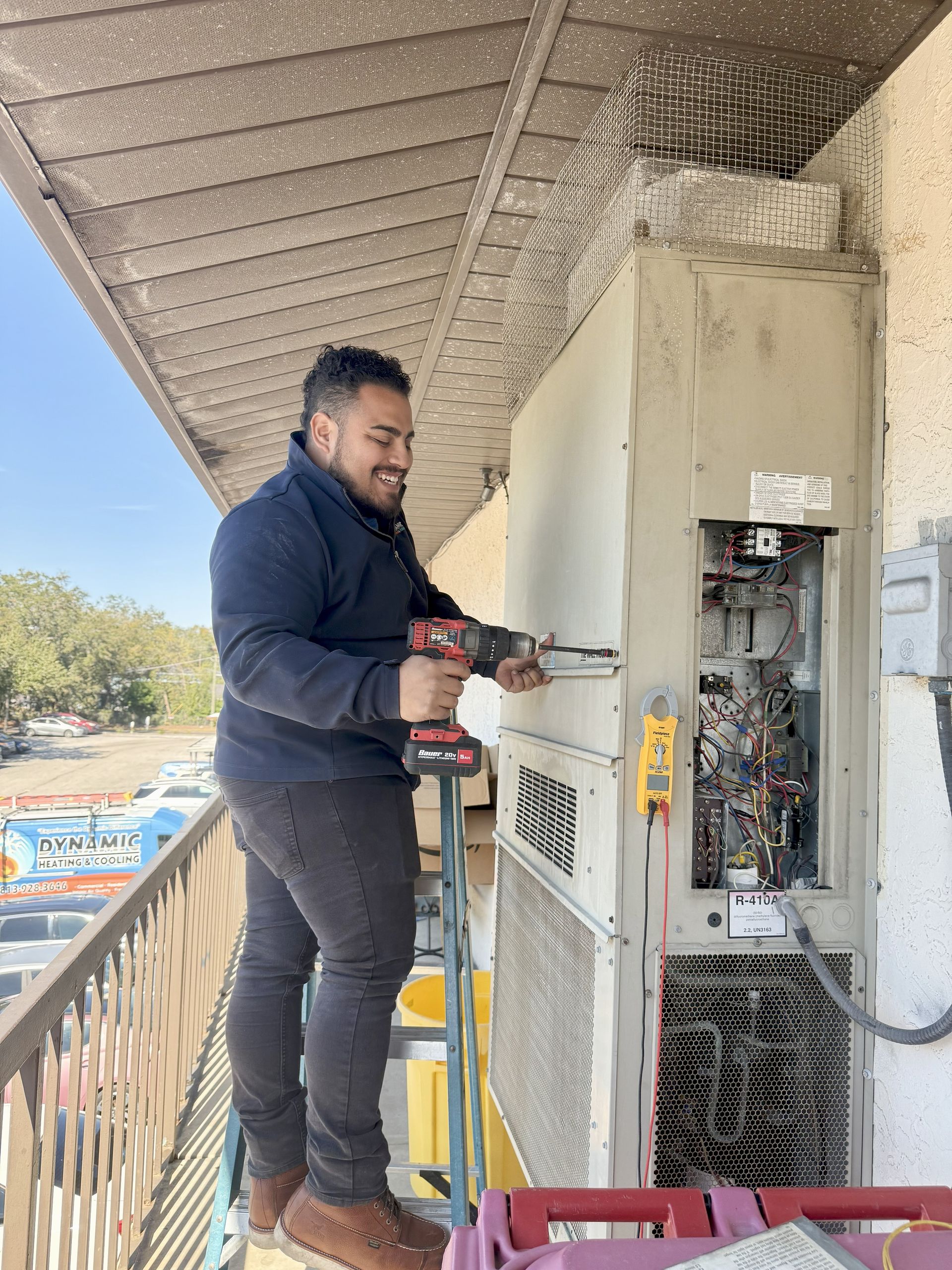 Man in dark clothing uses a power drill on a large outdoor HVAC unit on a balcony. He smiles while standing on a ladder.