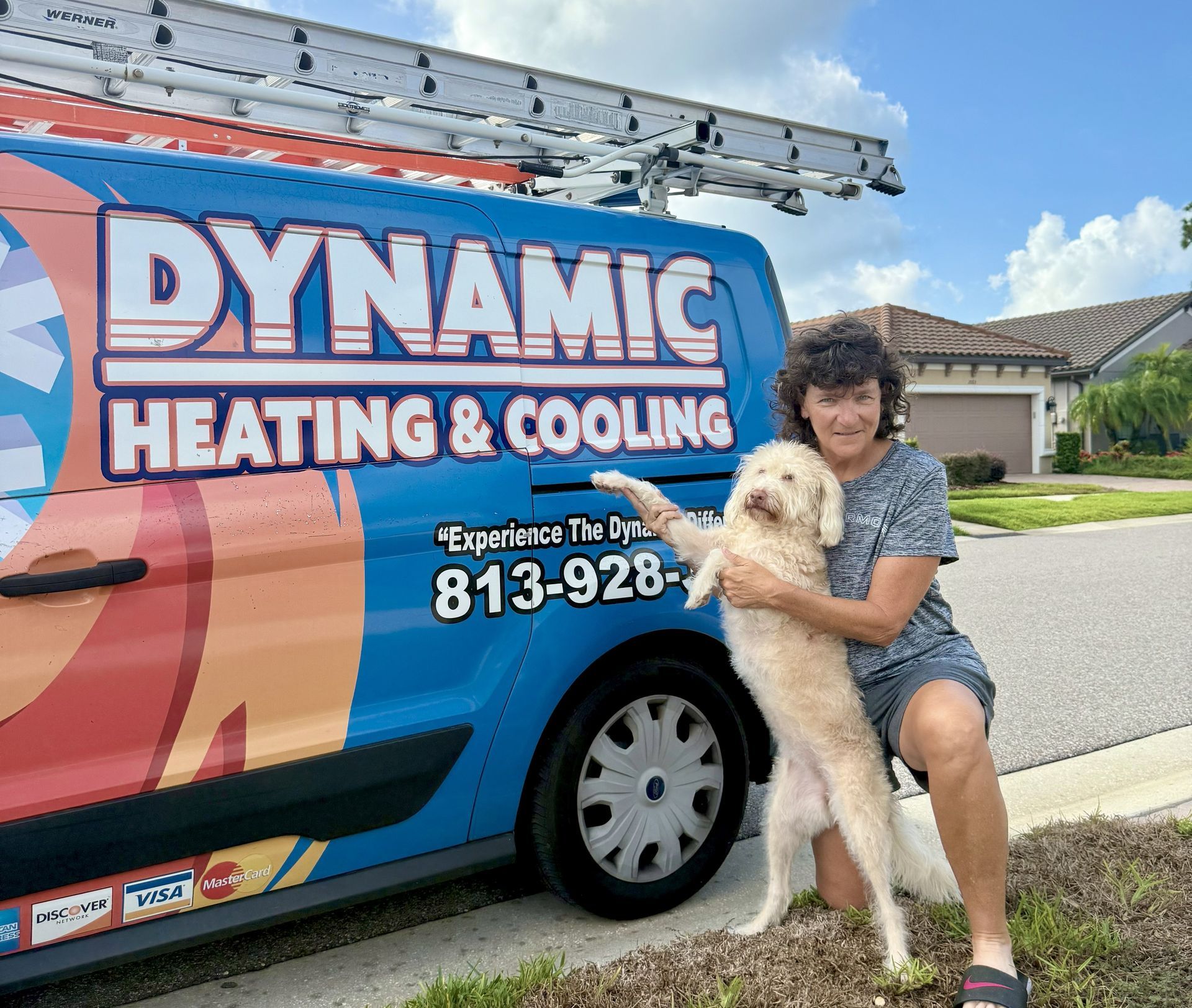 Woman kneels with a dog next to a Dynamic Heating & Cooling van in a residential setting. The van is blue with company logos.