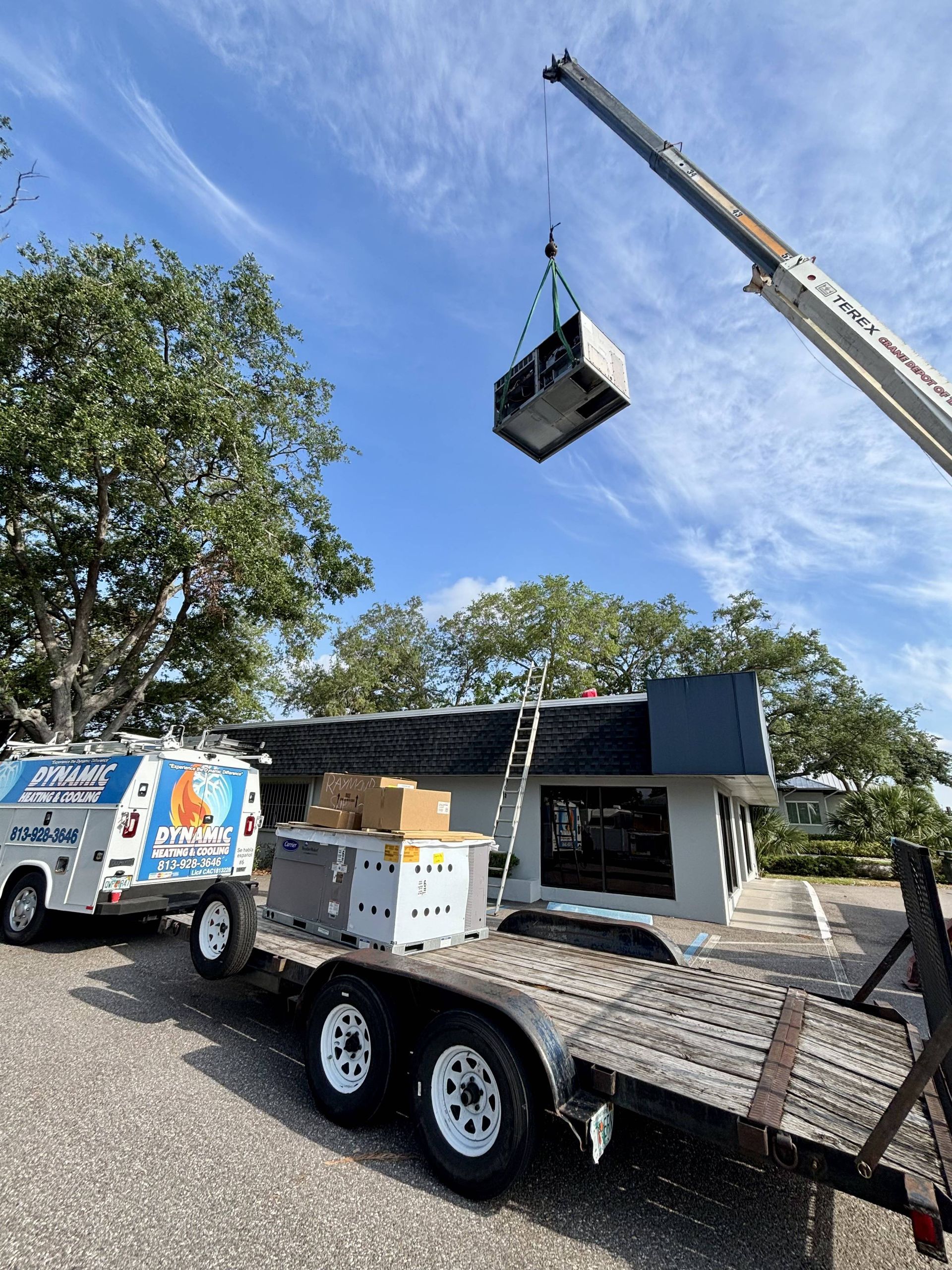 A crane lifts an AC unit to a building roof. A trailer, truck, and trees surround the building.