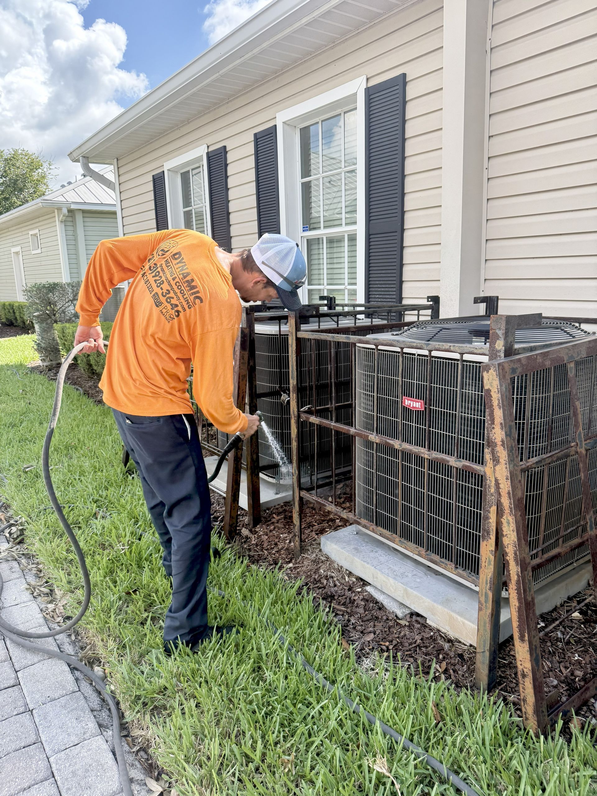 Man in orange shirt spraying an air conditioning unit outdoors. He is near a beige house with black shutters.