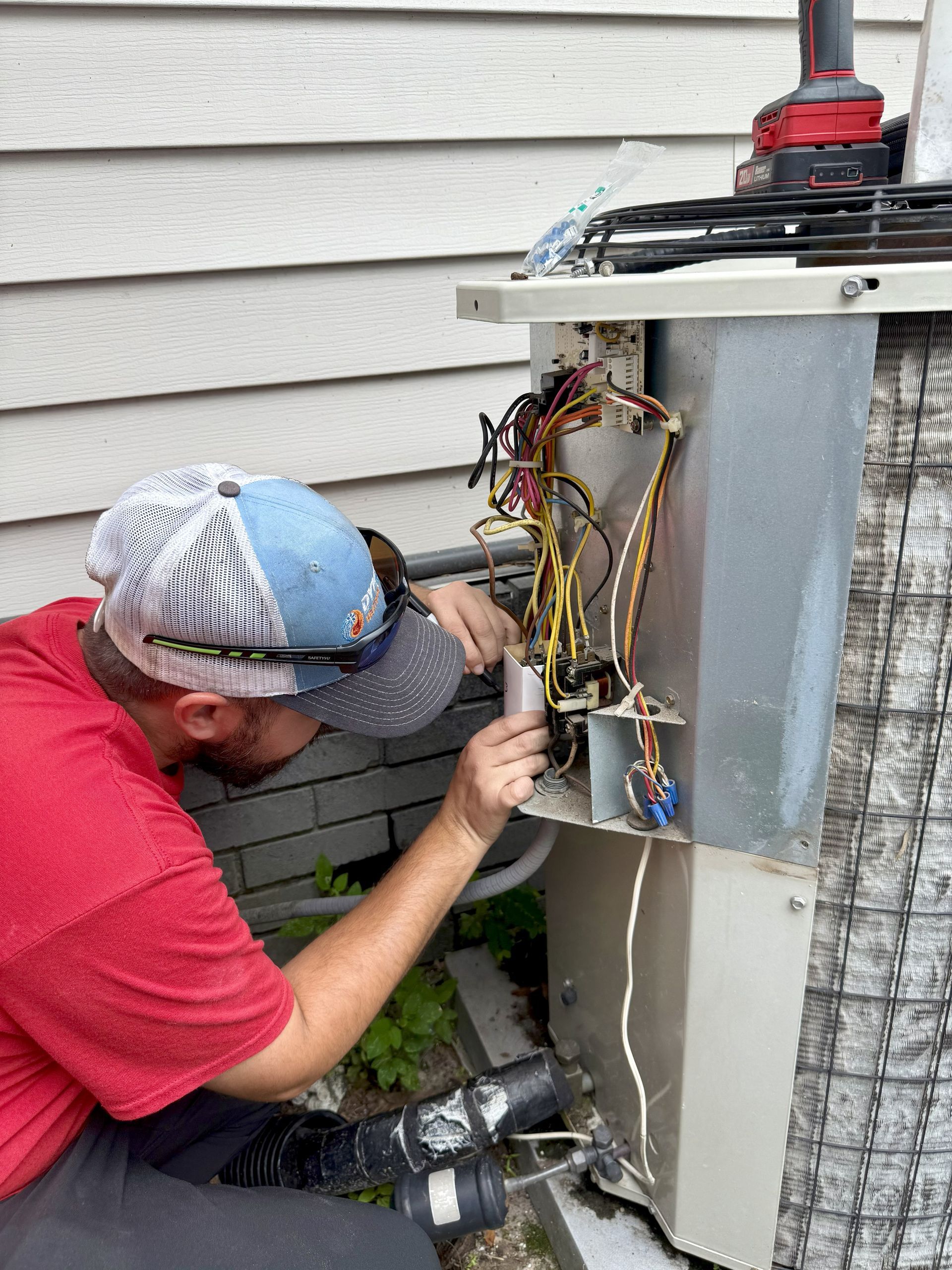 A man in a red shirt and baseball cap repairs an air conditioning unit, working with wires near a siding wall.