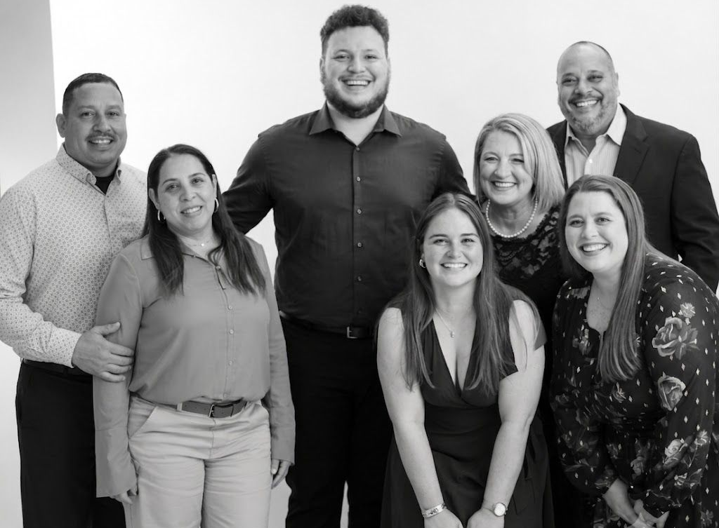 Seven smiling coworkers posed together in front of a plain backdrop.