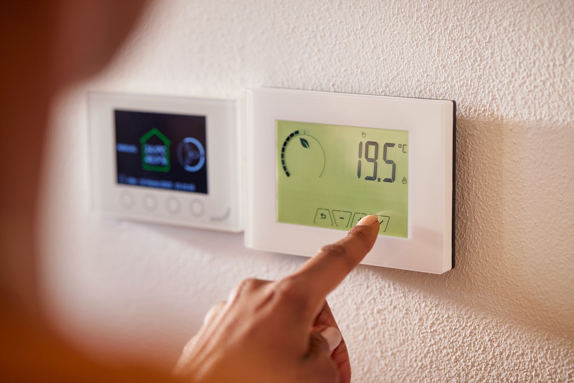 A hand adjusting a digital thermostat on a white wall, displaying a temperature of 19.5 degrees Celsius.