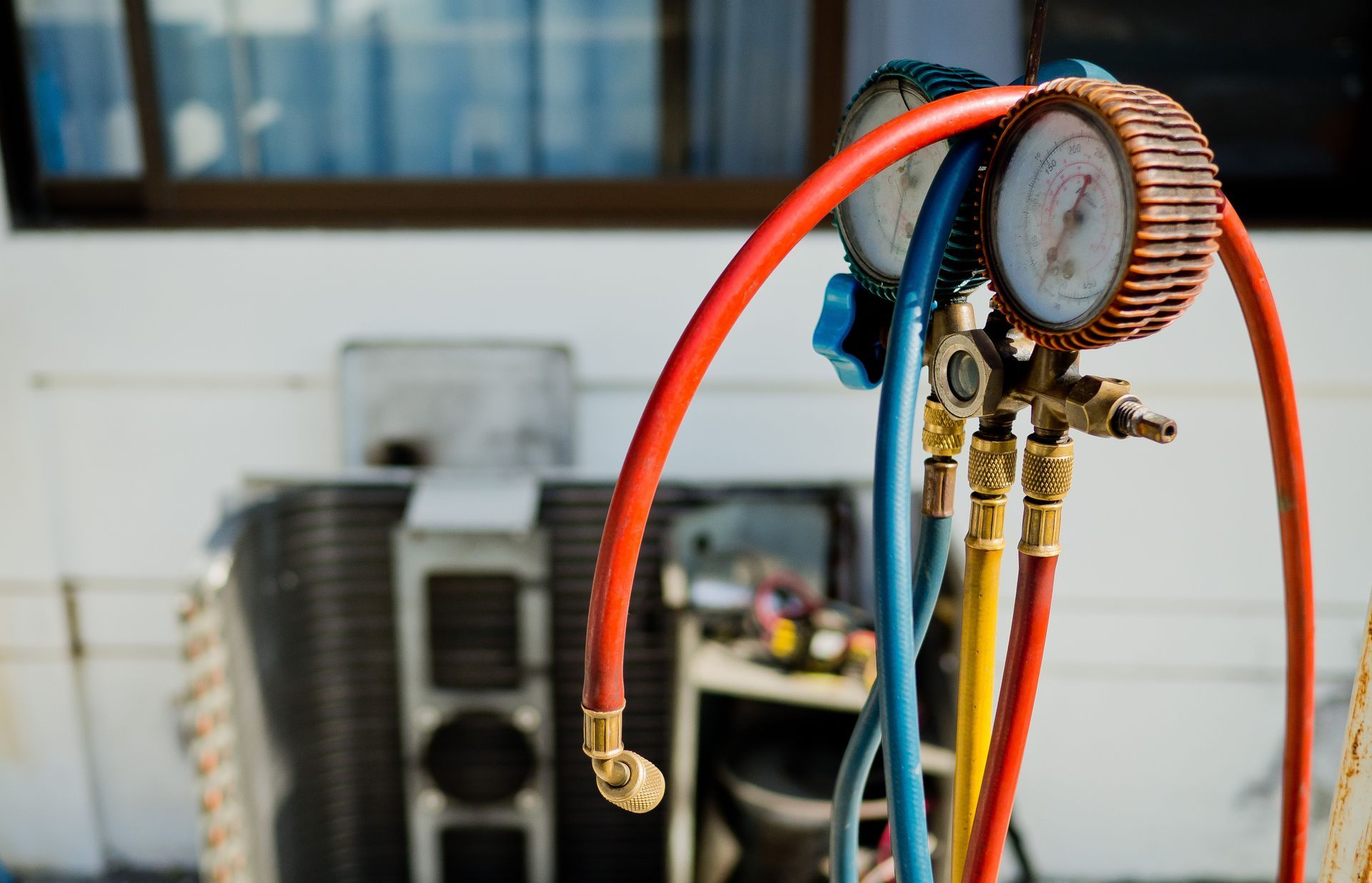 Close-up of pressure gauges with red, blue, and yellow hoses connected to an AC unit outside a building.