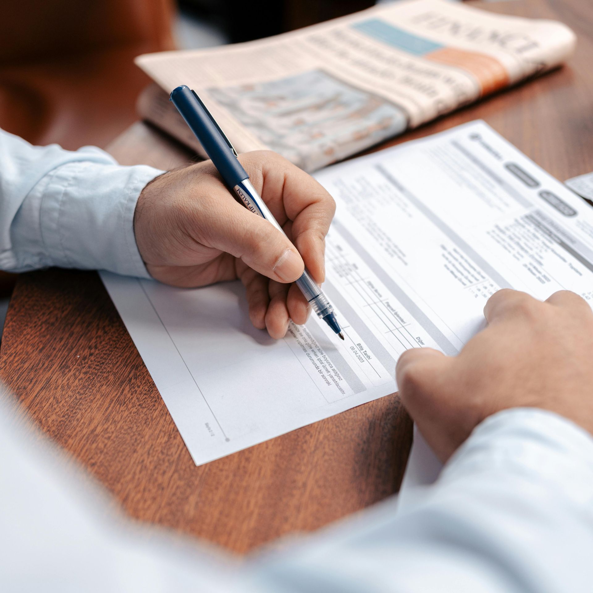 Hands signing a document with a pen on a wooden table, with paperwork and a newspaper nearby